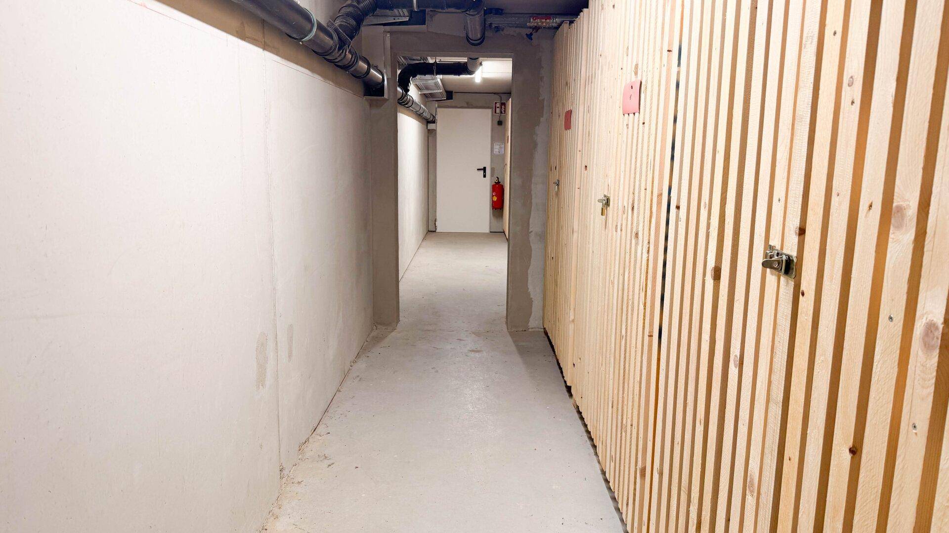 Heller Kellerflur mit individuellen Lagerabteilen aus Holzlatten und sichtbaren Rohren an der Decke.
