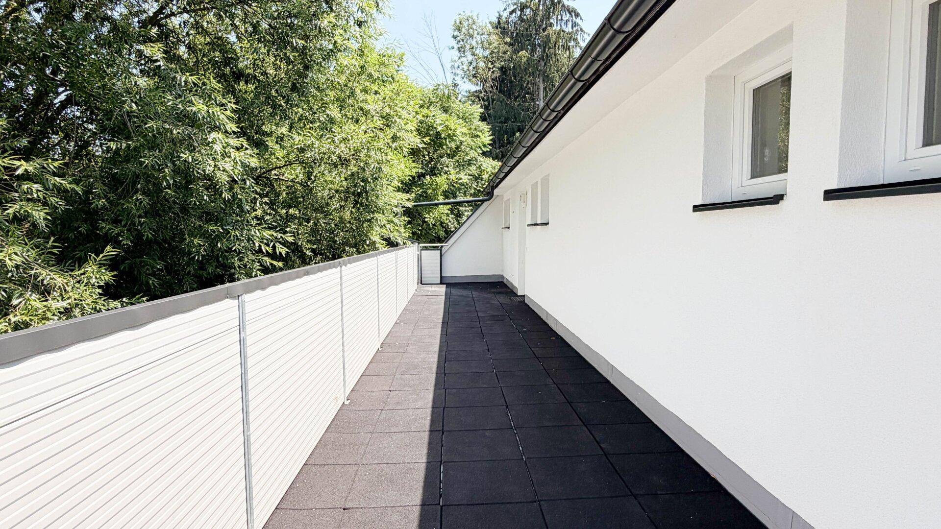 Langer Balkon mit dunklen Fliesen und grüner Aussicht, ideal für Entspannung im Freien.