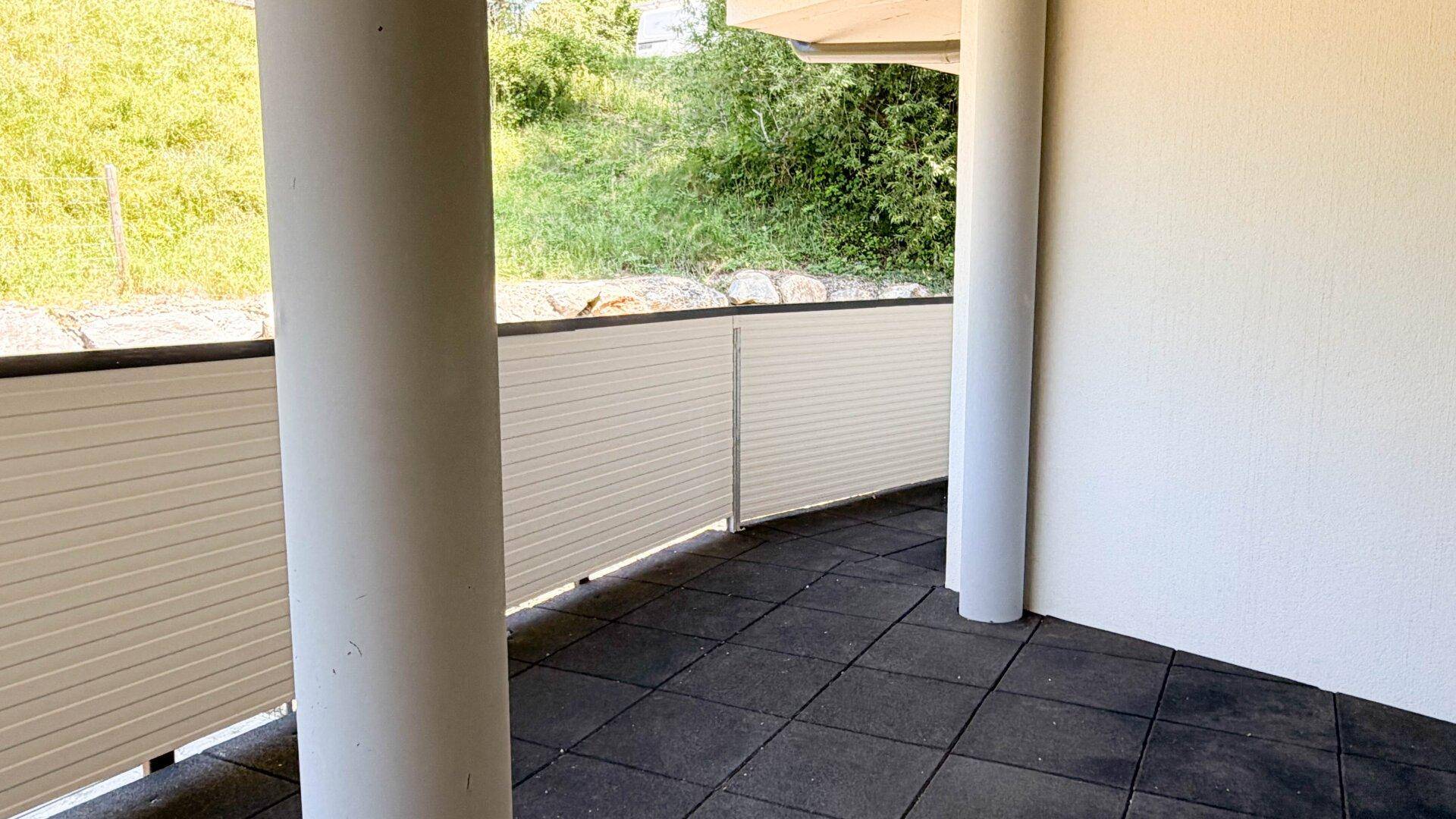 Balkon mit dunklem Fliesenboden und einer hellen Brüstung, bietet einen Blick ins Grüne.