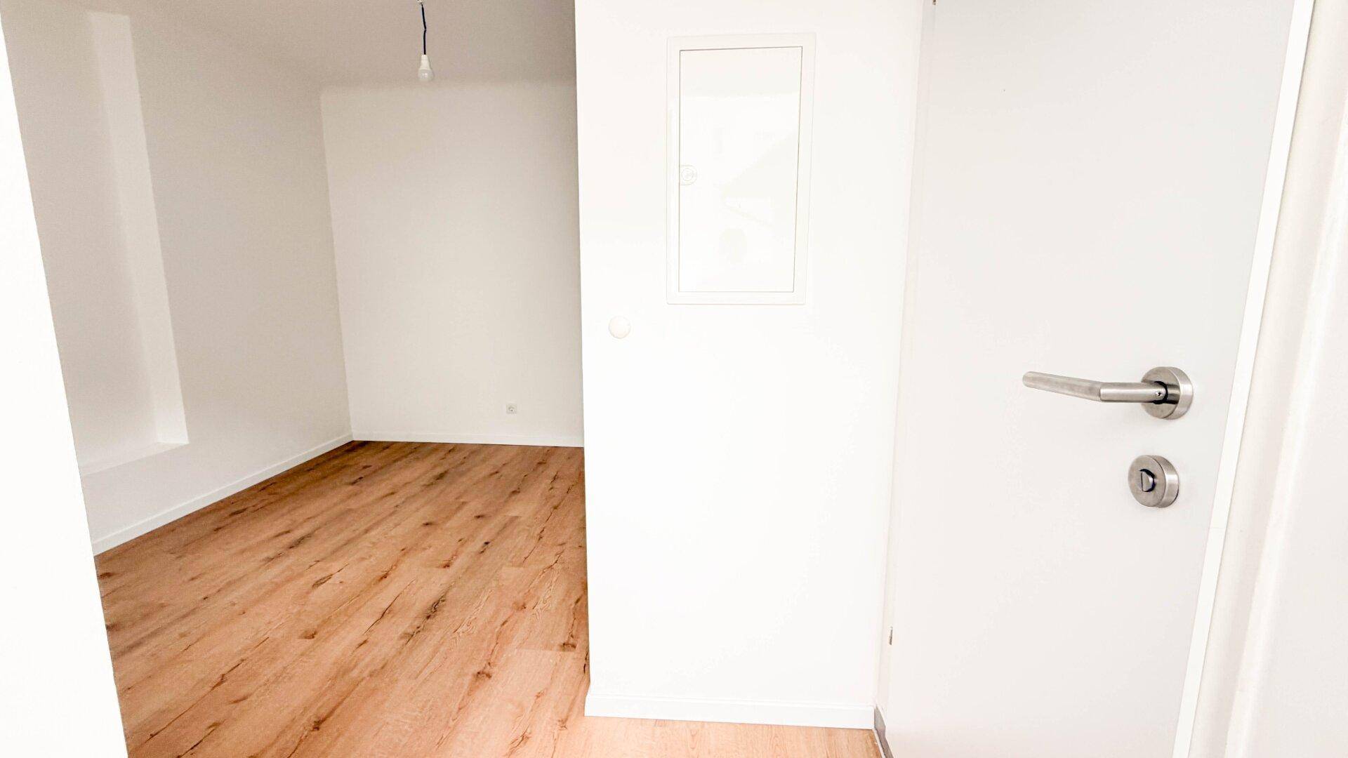 Heller Flur mit weißer Tür und Blick in einen angrenzenden Raum mit Holzfußboden.