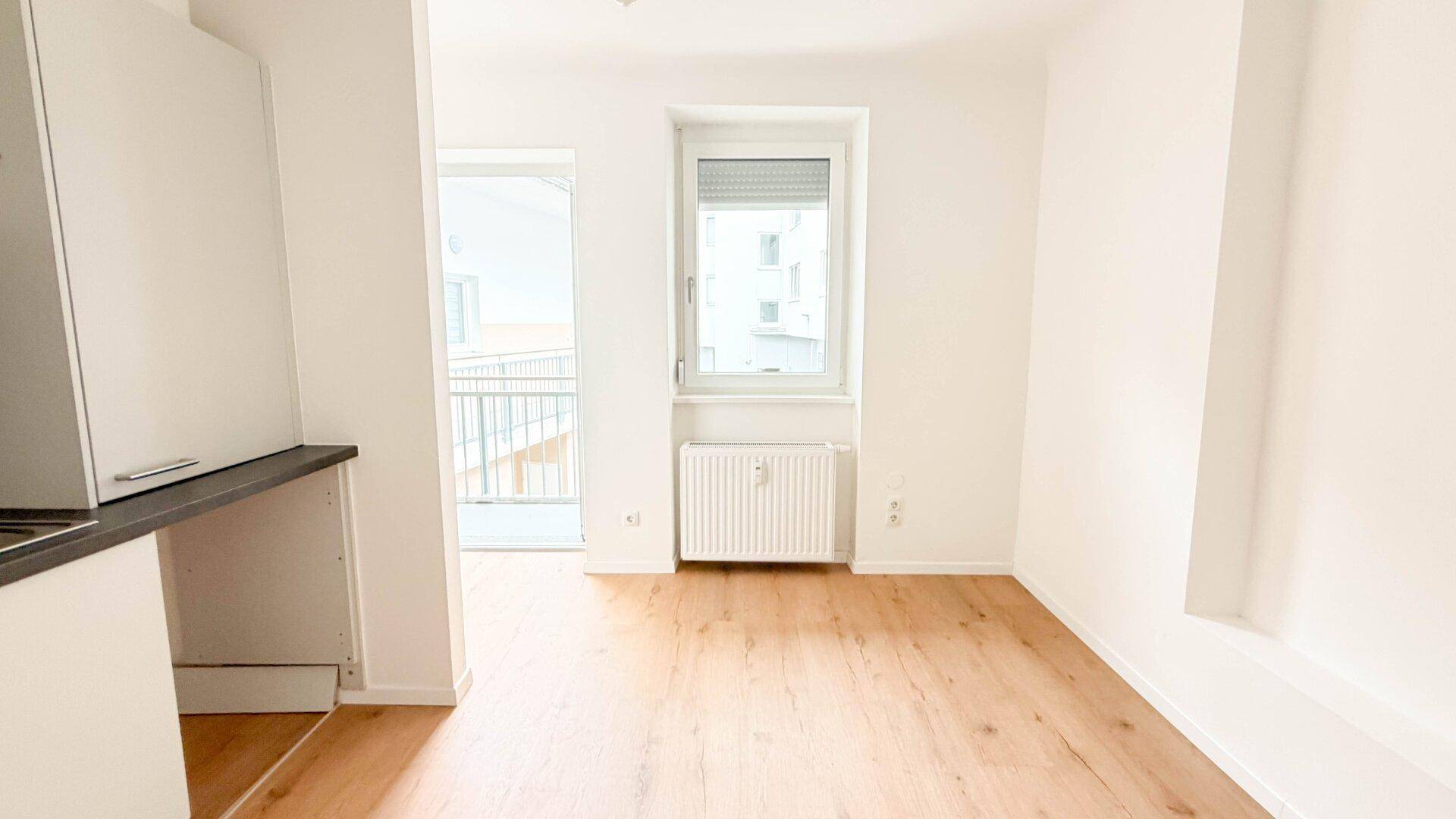 Offener Wohnbereich mit Küchenzeile, hellem Holzfußboden und Zugang zum Balkon.