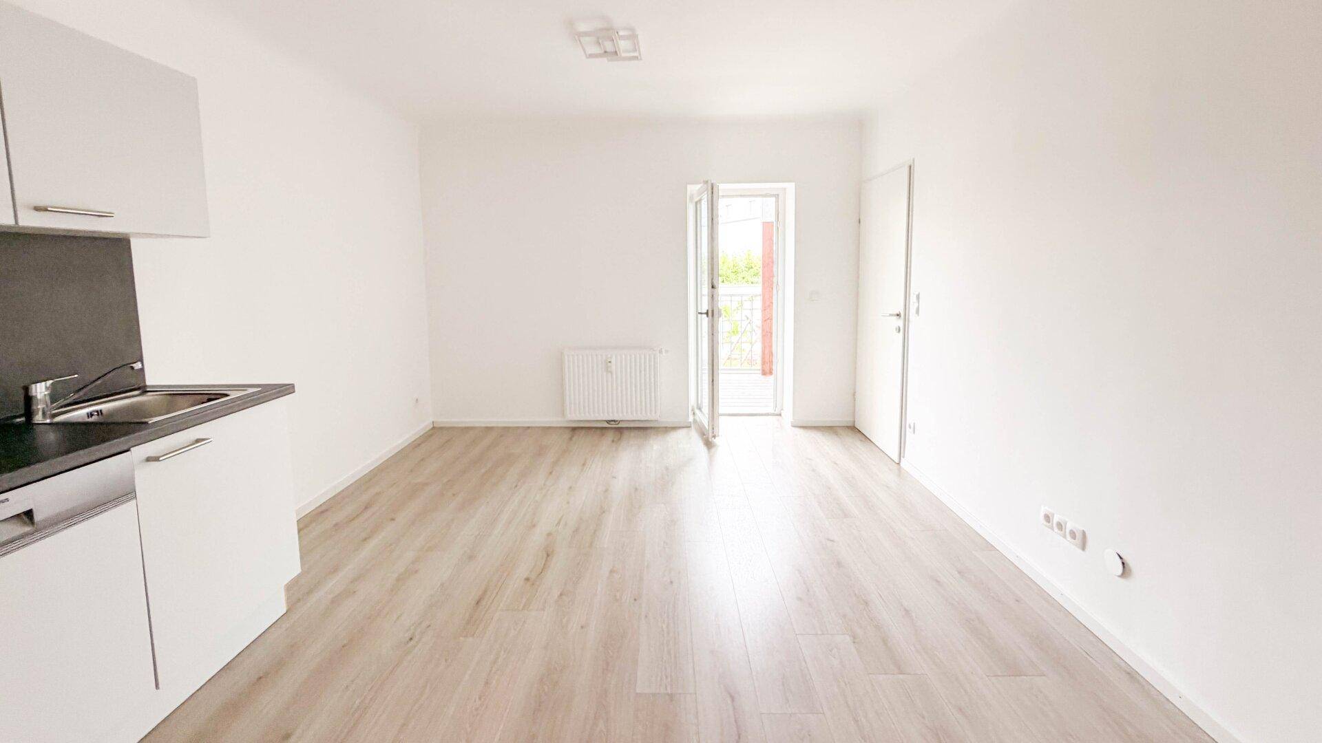 Heller Wohnraum mit Laminatboden und Zugang zu einem Balkon, ausgestattet mit einer kleinen Küchenzeile.