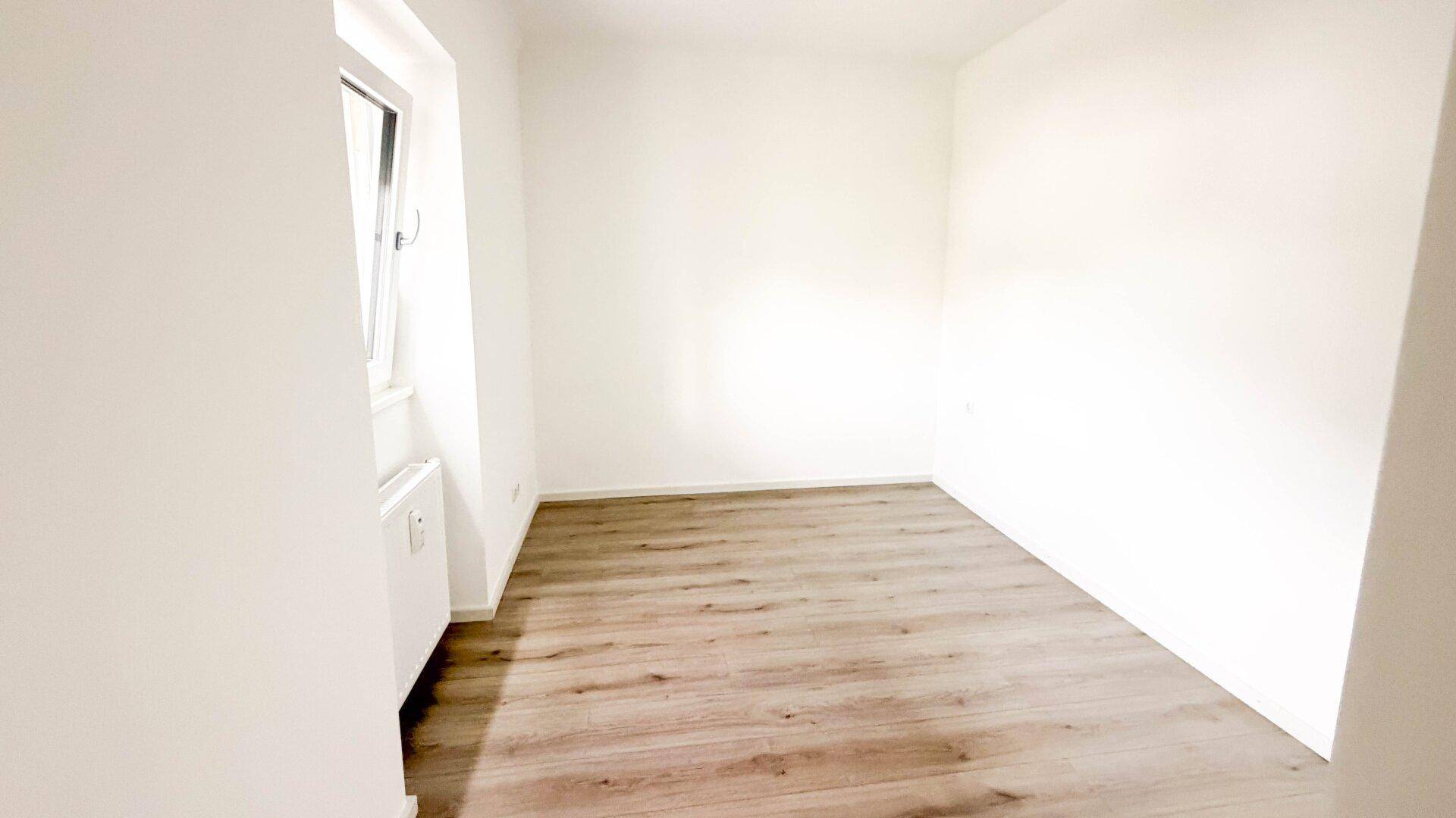 Helles Schlafzimmer mit Laminatboden und einem Fenster, das viel Tageslicht hereinlässt.