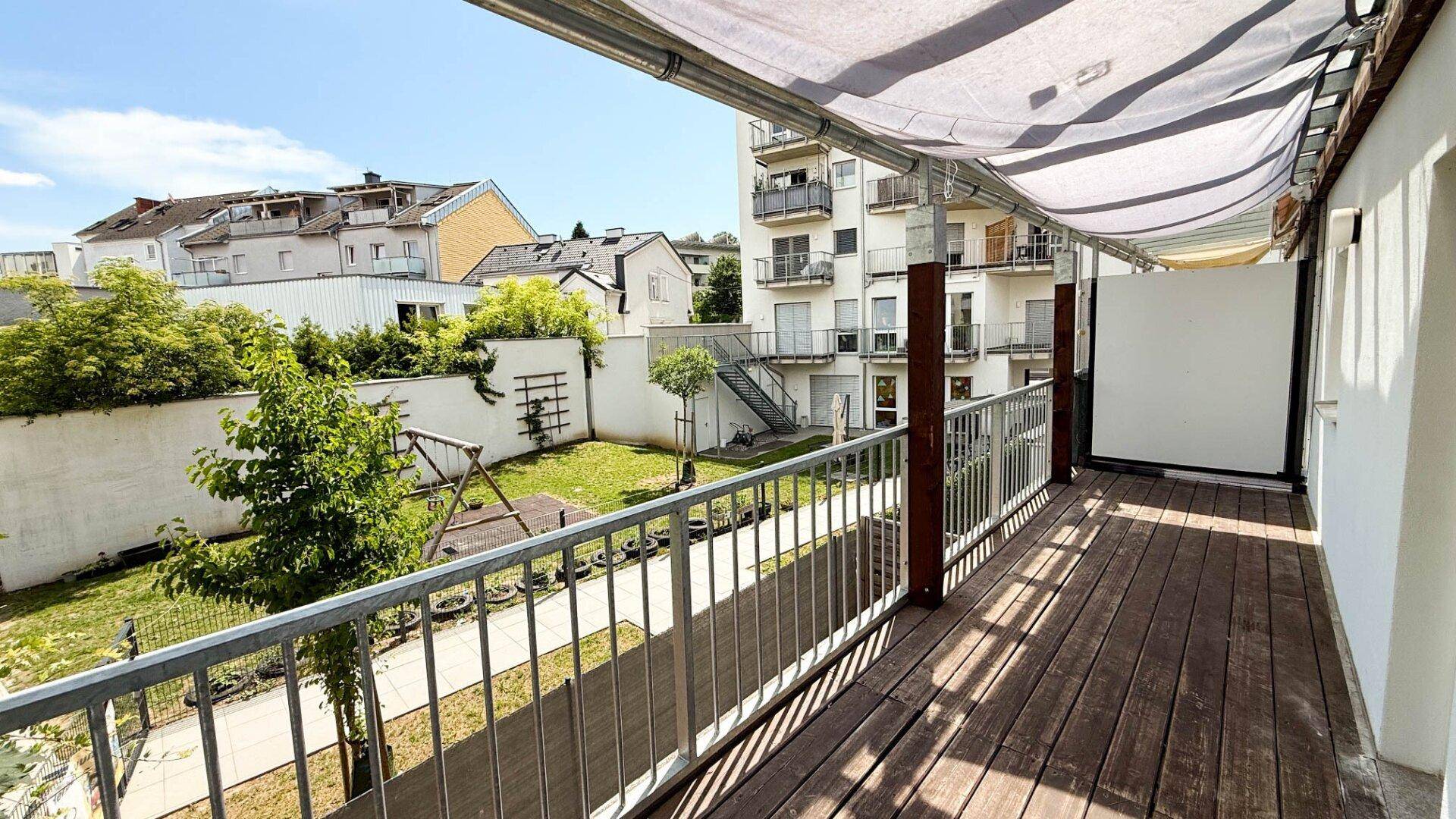 Langer Balkon mit Holzboden und Markise, bietet Blick auf den grünen Innenhof.