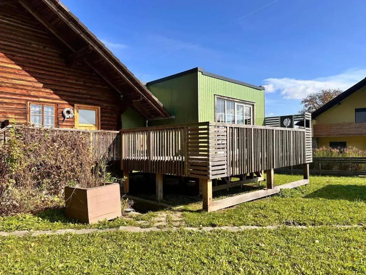 Detailansicht der Holzterrasse am Blockhaus mit grünem Anbau unter strahlendem Himmel.