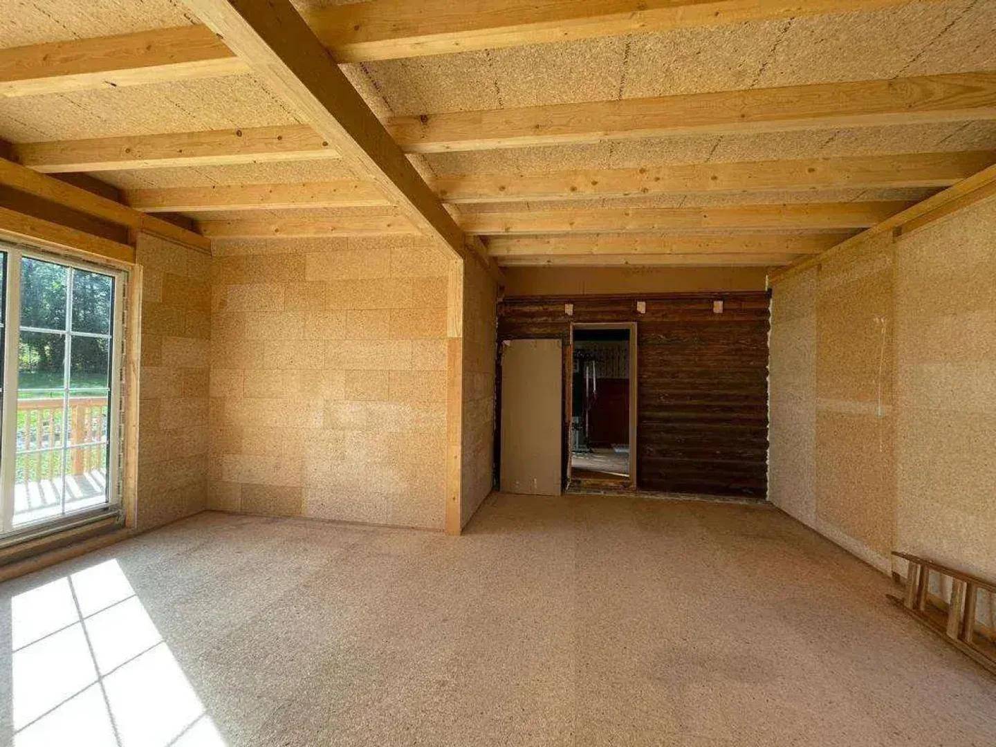 Unfertiger Innenraum mit Holzbalkendecke und Zugang zur Terrasse, viel natürliches Licht.