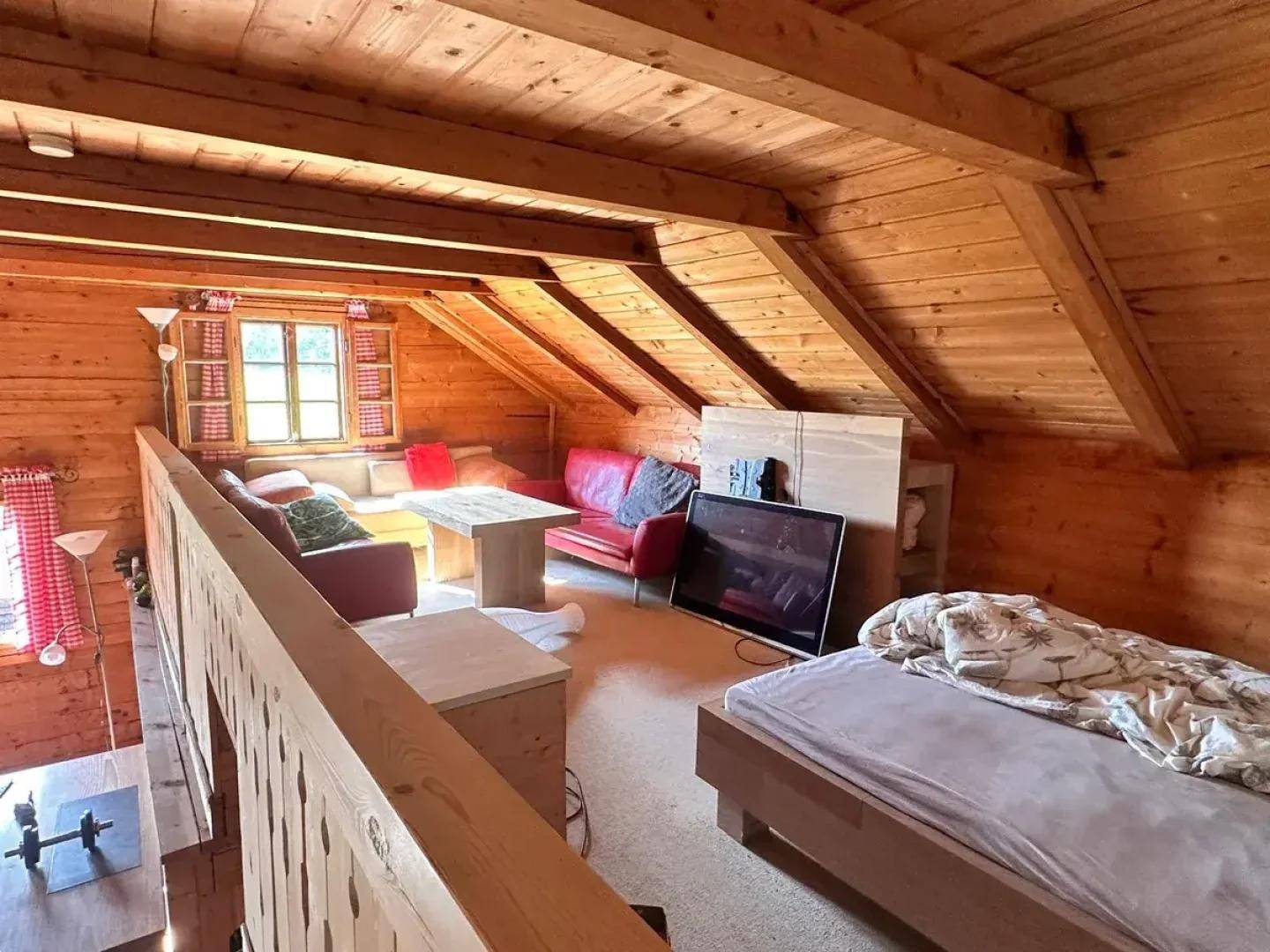 Rustikaler Wohnbereich mit Holzwänden, Holzbalkendecke und Schlafgelegenheit im Dachgeschoss.