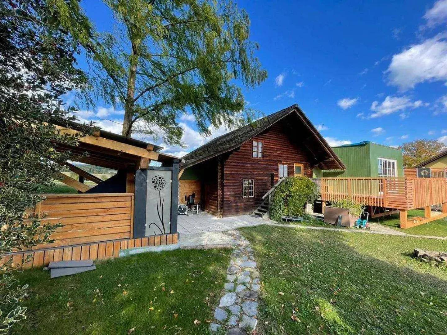 Blockhaus mit modernem Anbau, Holzterrasse und überdachtem Unterstand im Grünen.