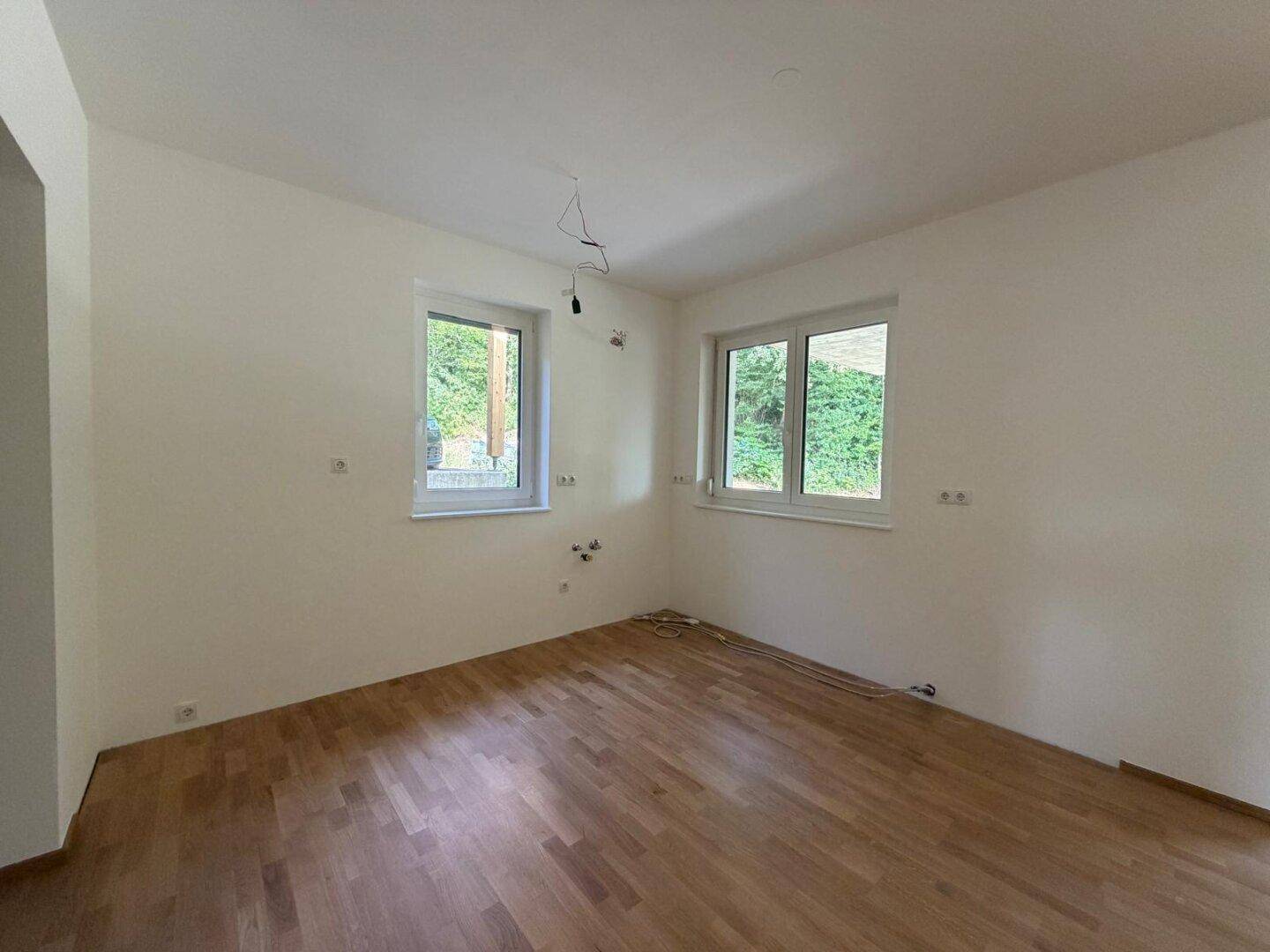 Leeres Zimmer mit Holzboden, weißen Wänden und zwei Fenstern mit Blick ins Grüne.