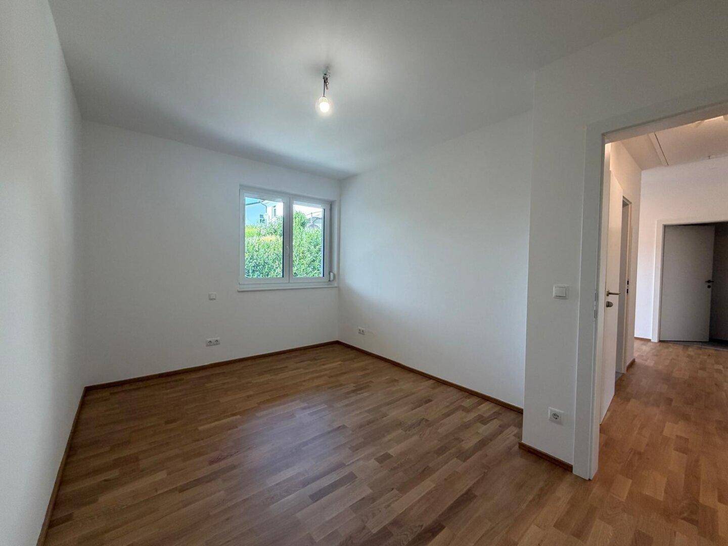 Leeres Schlafzimmer mit Holzboden, weißen Wänden und Fenster mit Grünblick.