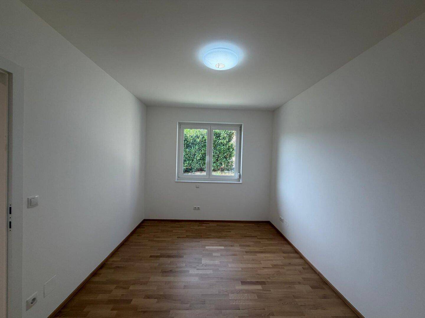 Helles Zimmer mit Holzboden, weißen Wänden und Fenster mit Blick ins Grüne.