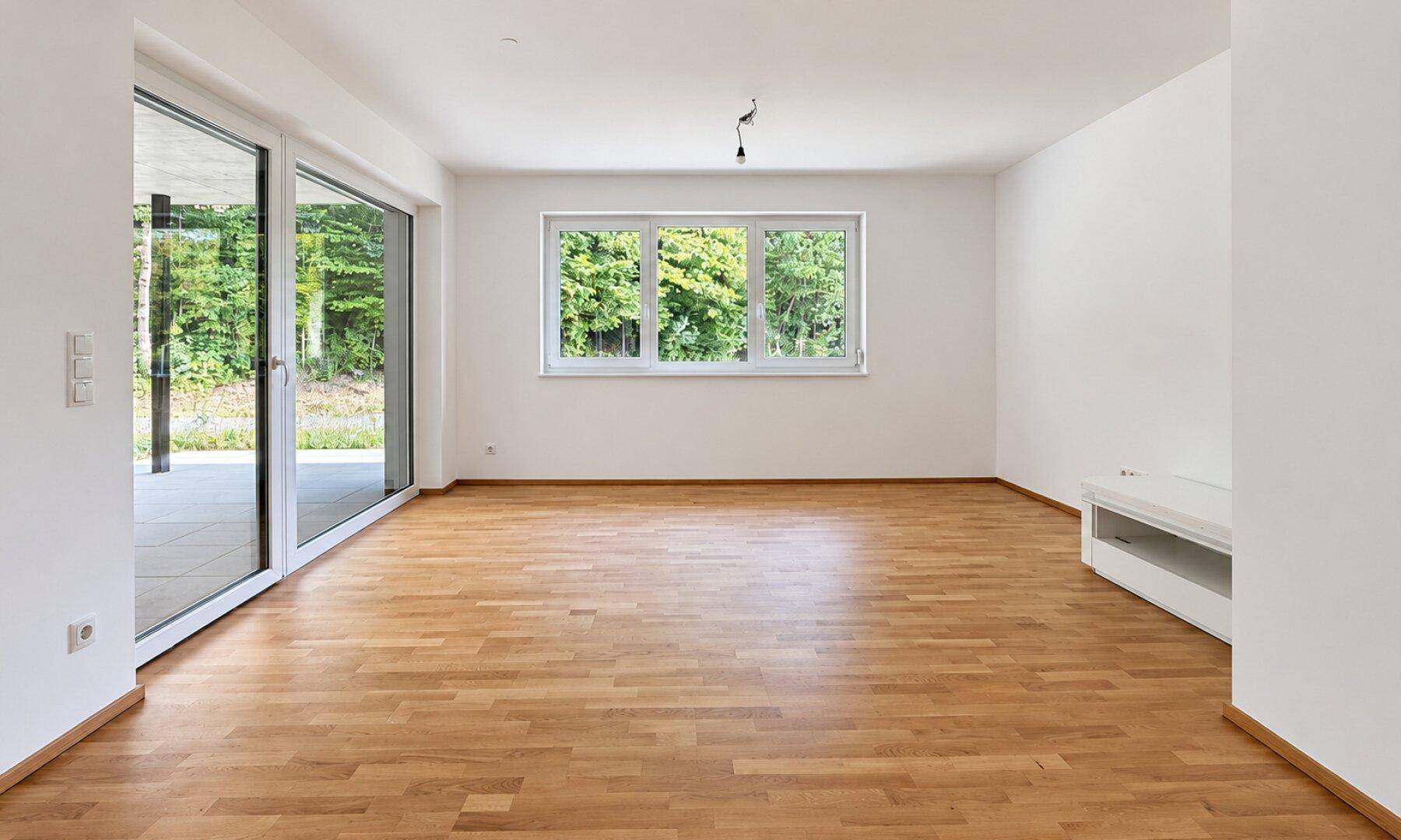 Helles, leeres Wohnzimmer mit großen Fenstern, Holzboden und Zugang zur Terrasse.