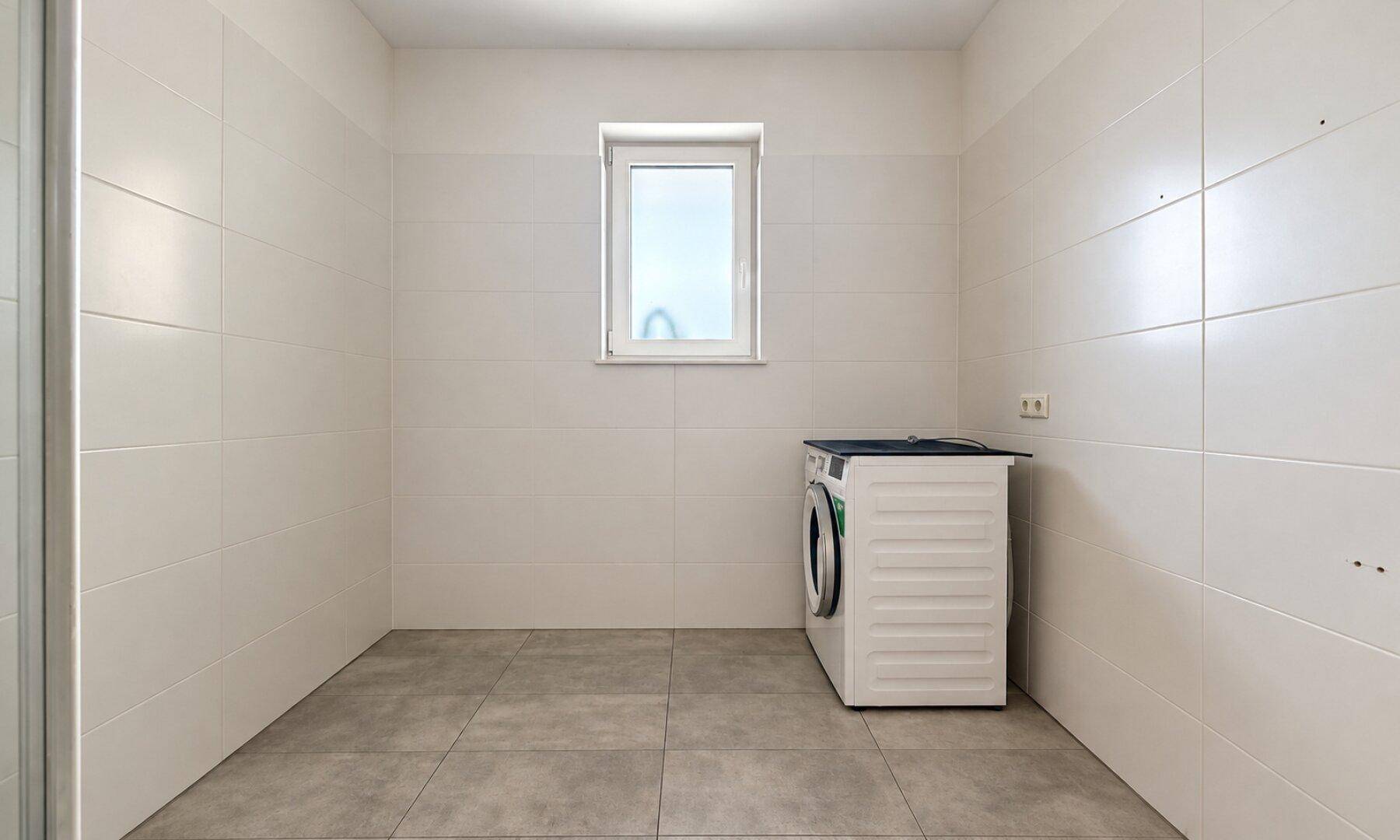 Praktischer Hauswirtschaftsraum mit Waschmaschine, Fliesenboden und Fenster für Belüftung.