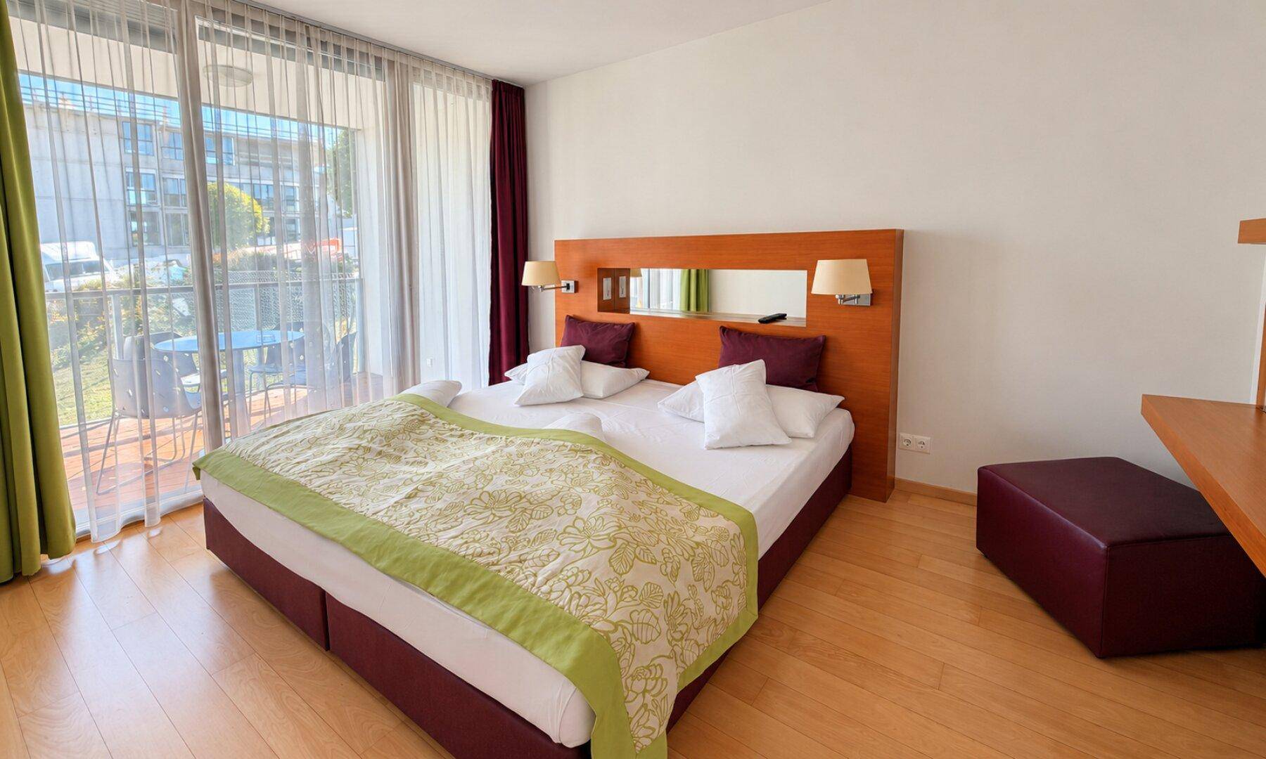Helles Schlafzimmer mit Doppelbett, Holzbettrahmen und Blick auf den Außenbereich.