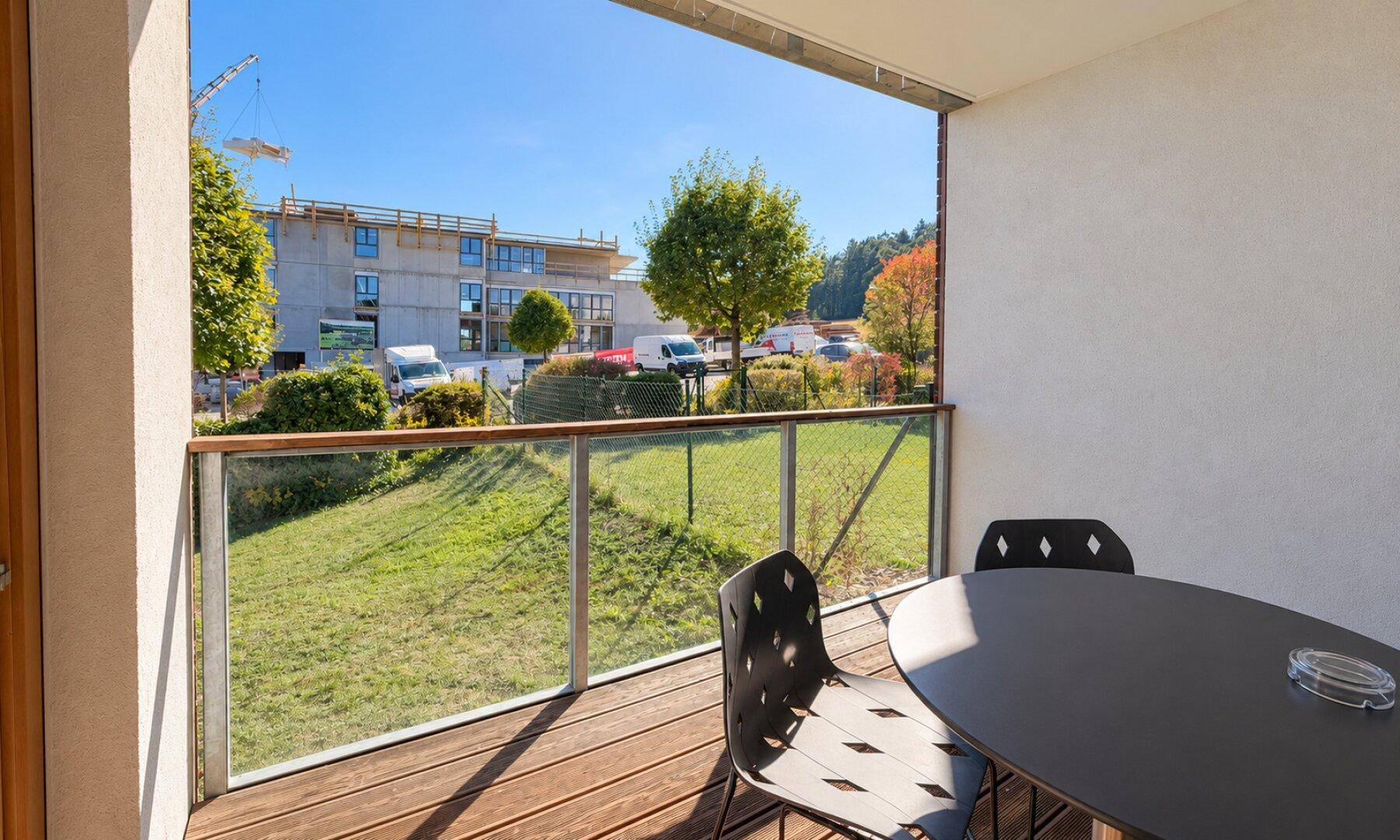 Geräumiger Balkon mit Holzdielen, Glasgeländer und Blick auf eine Baustelle und Grünflächen.