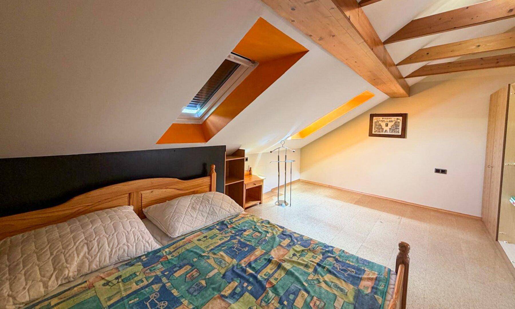 Dachgeschoss-Schlafzimmer mit Holzbalken, Dachfenster und orangefarbenen Akzenten.