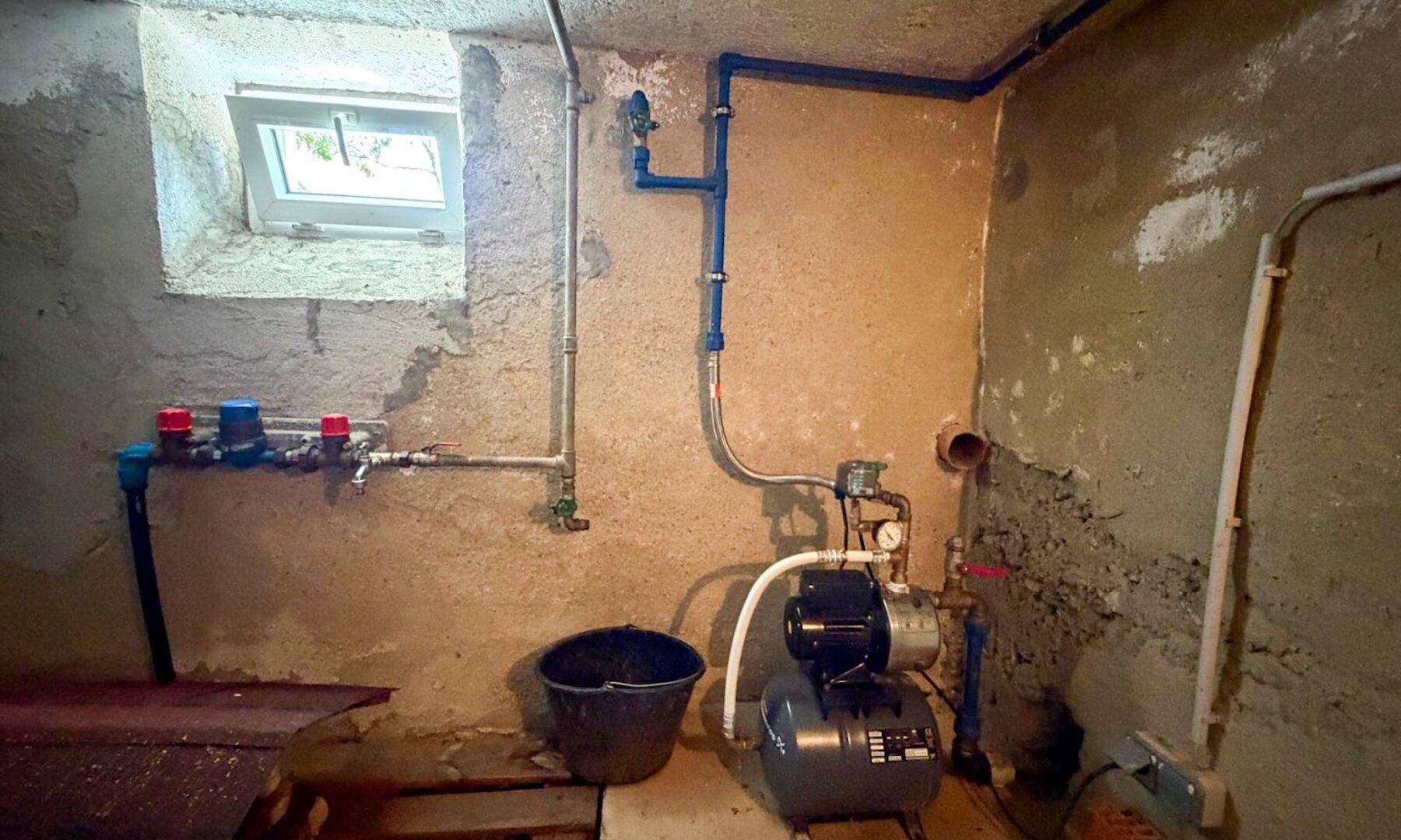 Kellerraum mit Wasserpumpe, sichtbaren Leitungen und kleinem Fenster.