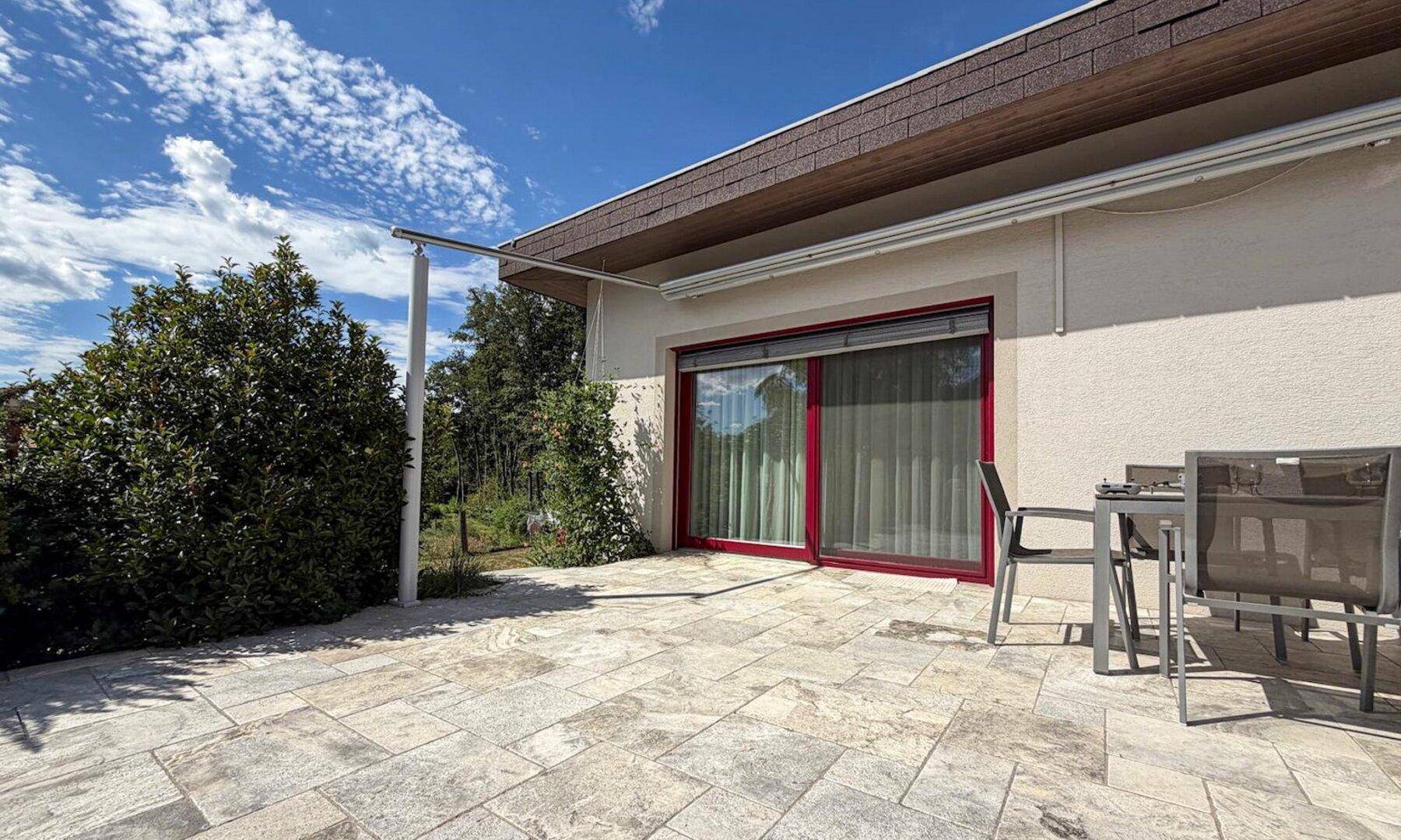 Große gepflasterte Terrasse mit Gartenmöbeln und Zugang zum Haus, umgeben von Grün.