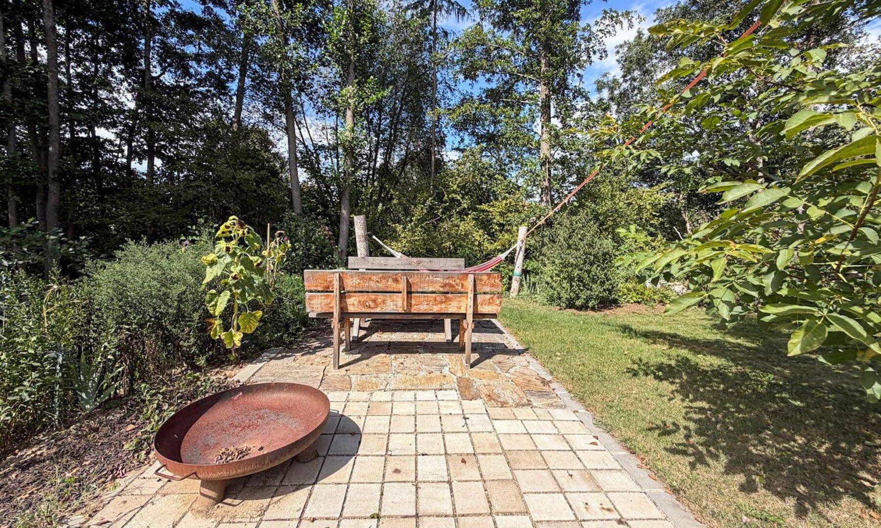 Idyllischer Gartenbereich mit Feuerstelle, Hochbeet und umgebenden Bäumen.