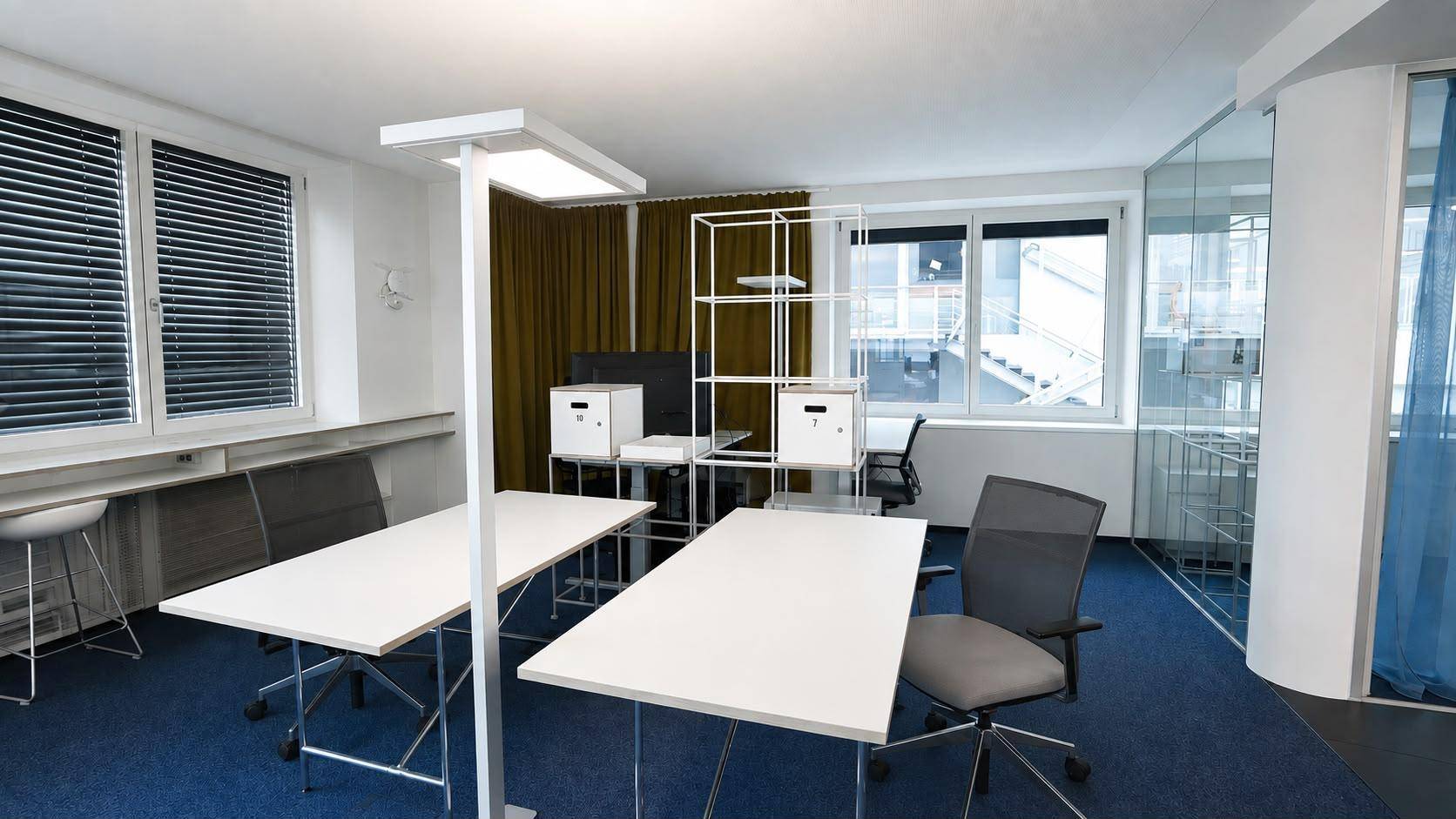 Funktionales Büro mit blauem Teppichboden, Schreibtischen und Fenstern mit Jalousien.