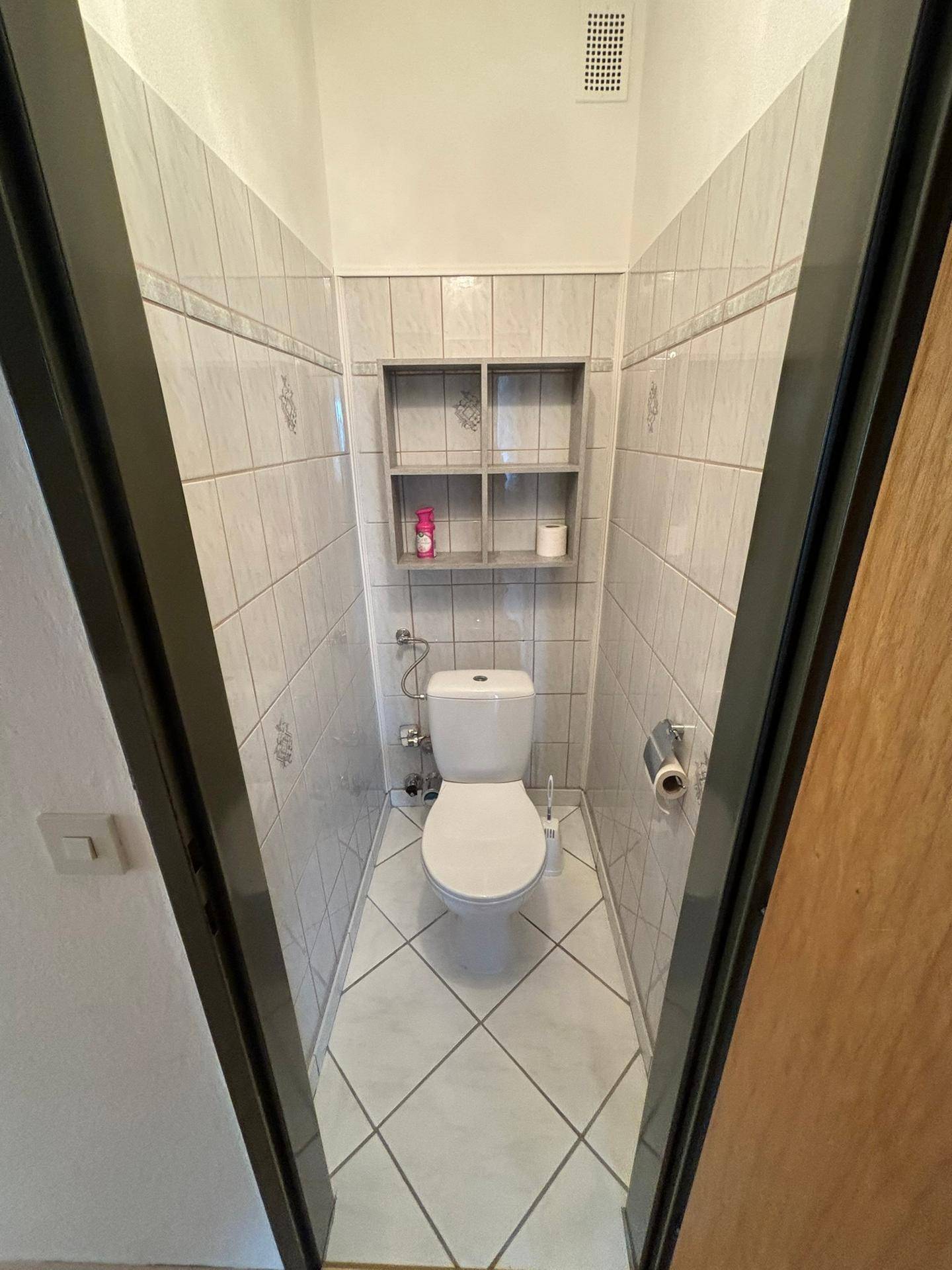 Separate Toilette mit weißen Fliesen und kleinem Regal, sauber und funktional.