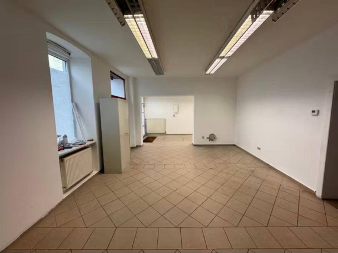 Geräumiger, leerer Raum mit Fliesenboden, weißen Wänden und großen Fenstern.
