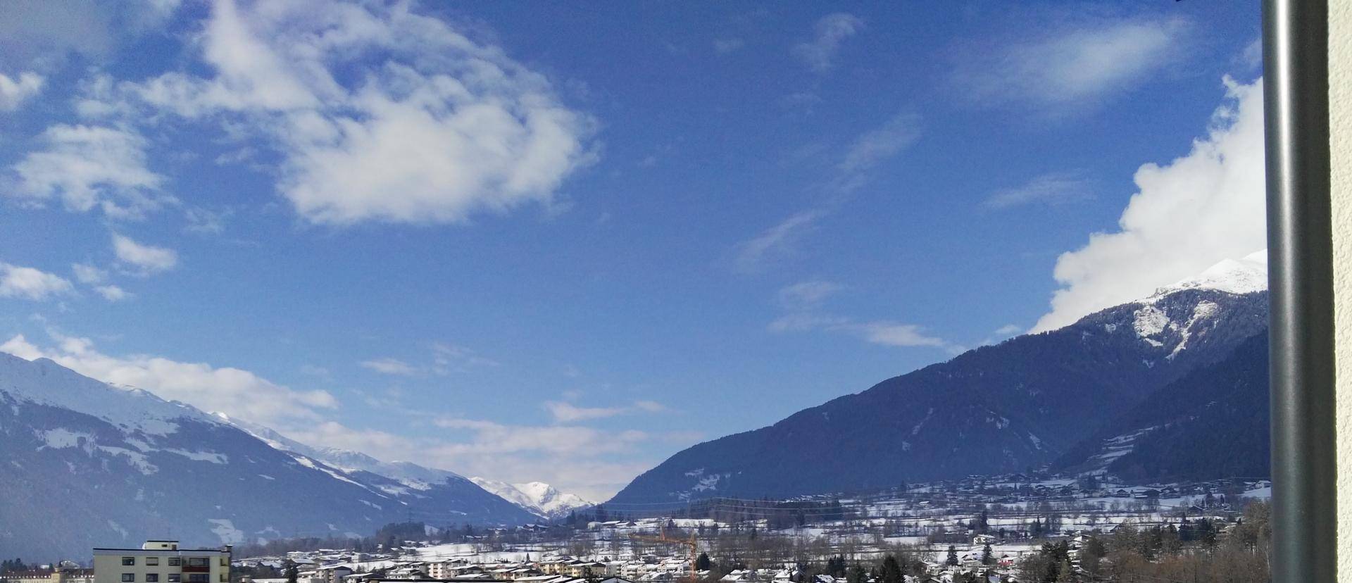 Weitblick auf schneebedeckte Berge und eine sonnige Stadtlandschaft unter blauem Himmel.