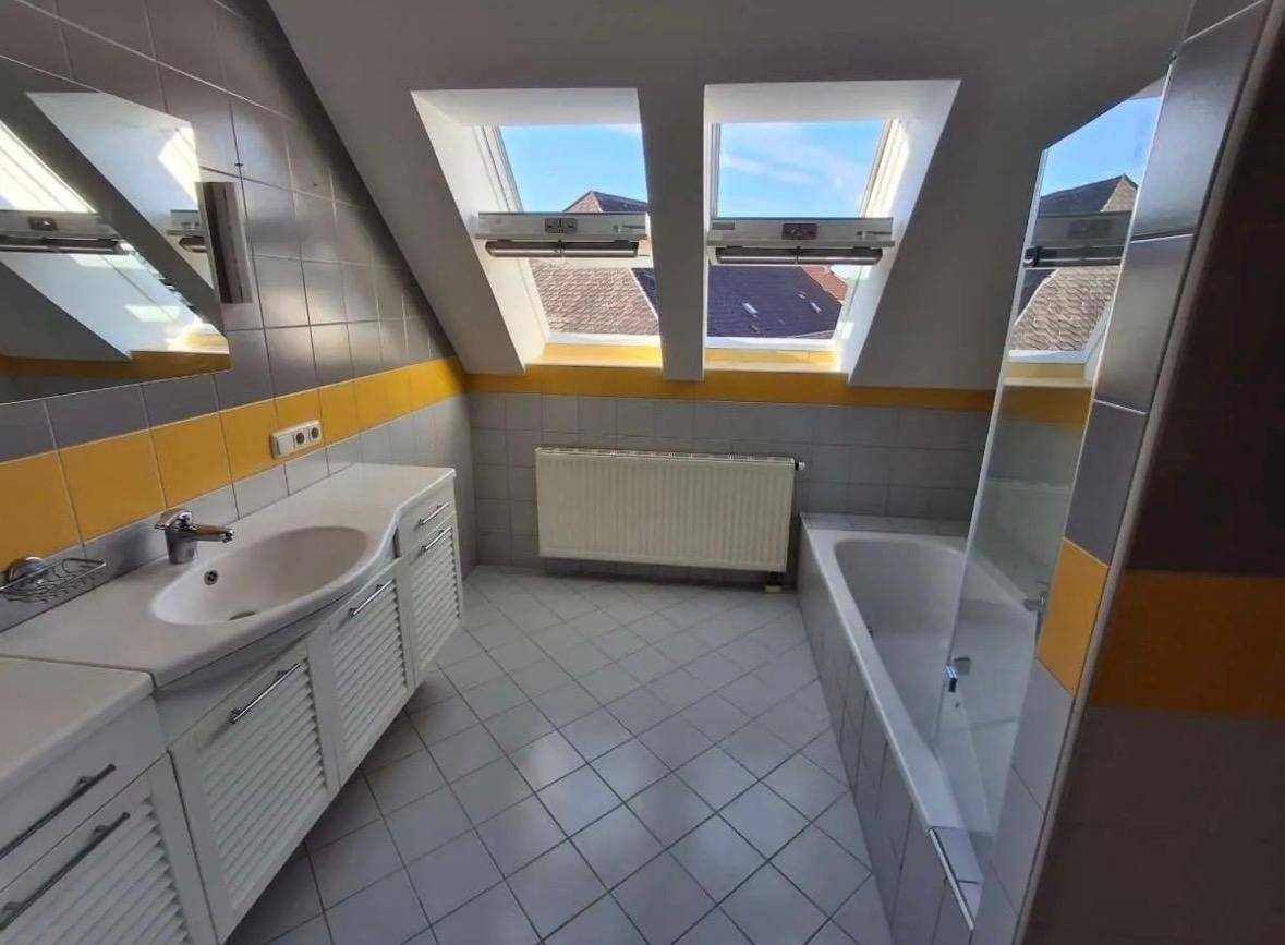 Helles Badezimmer mit Badewanne, Waschbecken und farbigen Fliesen unter zwei Dachfenstern.