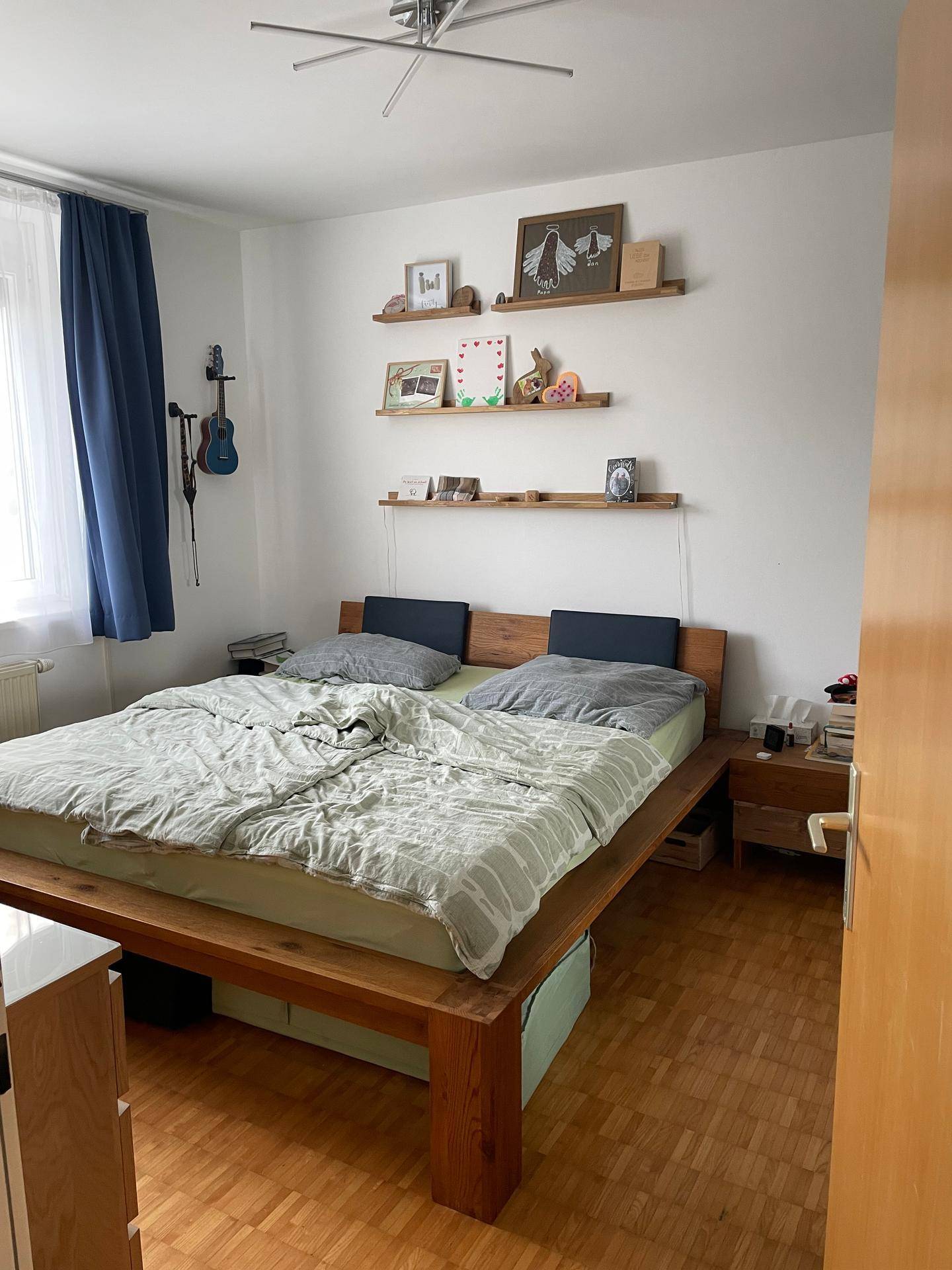 Gemütliches Schlafzimmer mit Holzbett, Parkettboden, blauen Vorhängen und dekorativen Wandregalen.