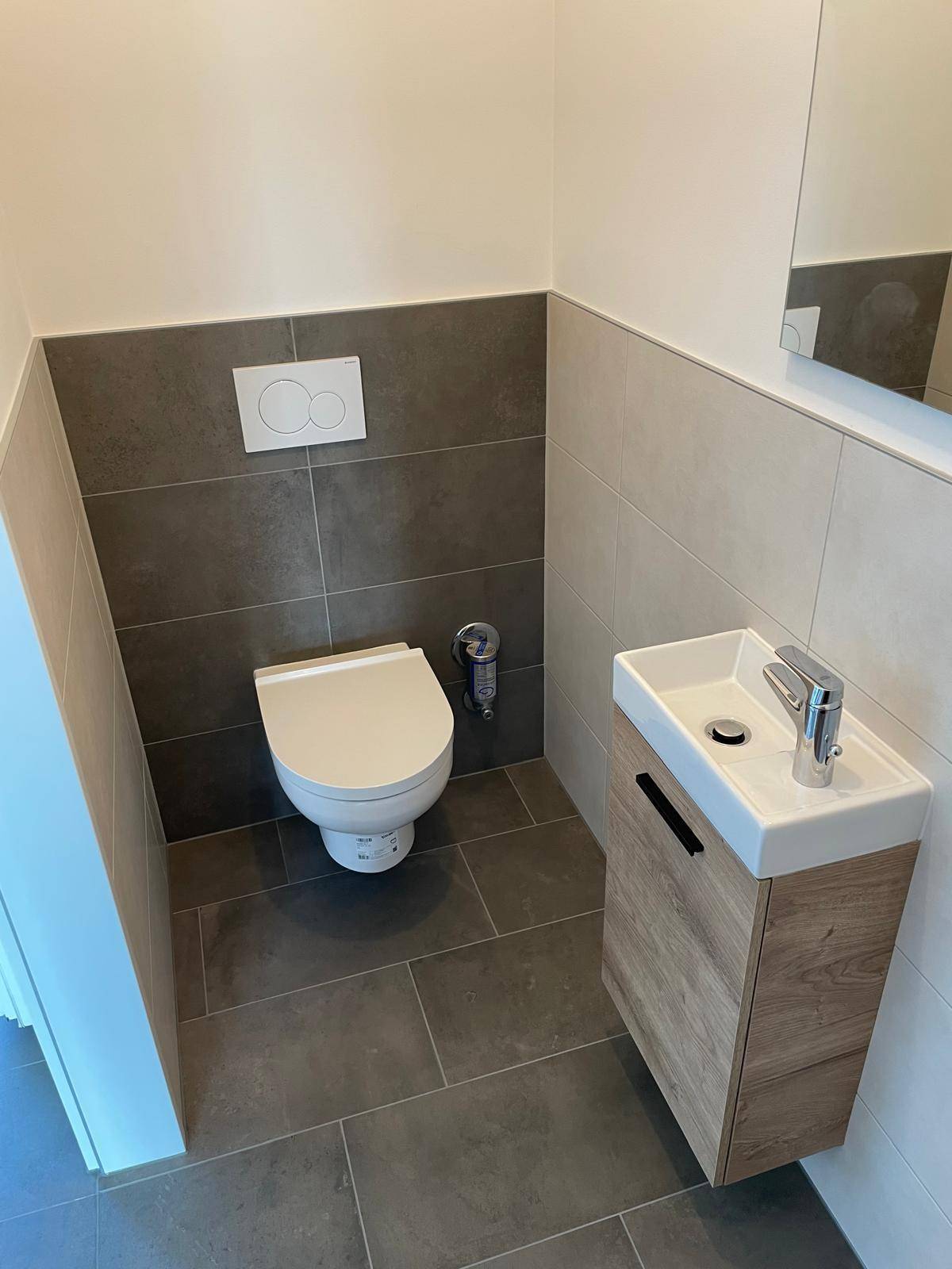 Modernes Gäste-WC mit wandhängender Toilette, kleinem Waschbecken und grauen Fliesen an Wänden und Boden.