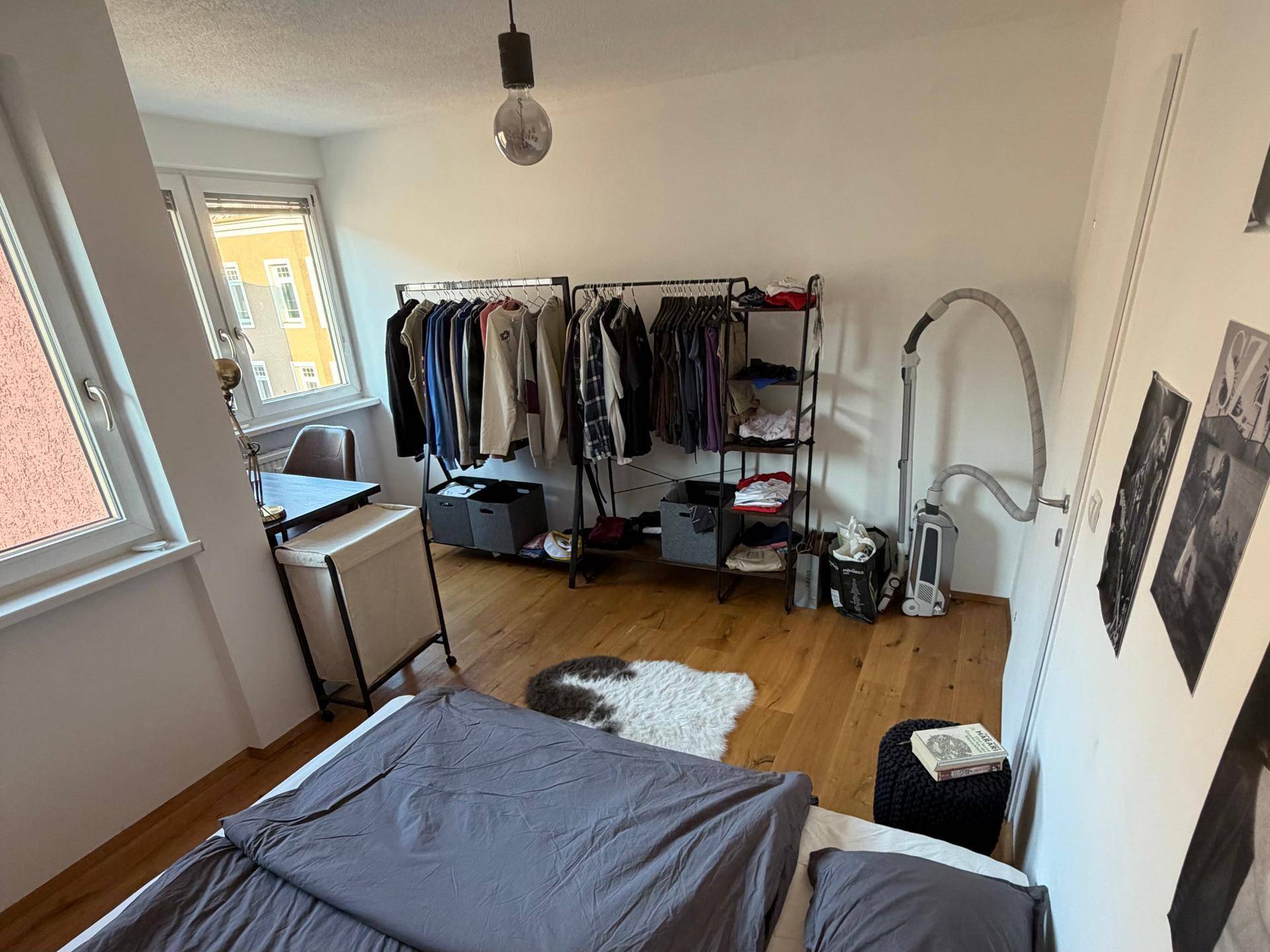 Schlafzimmer mit Holzboden, Kleiderständern und einem Schreibtisch am Fenster.