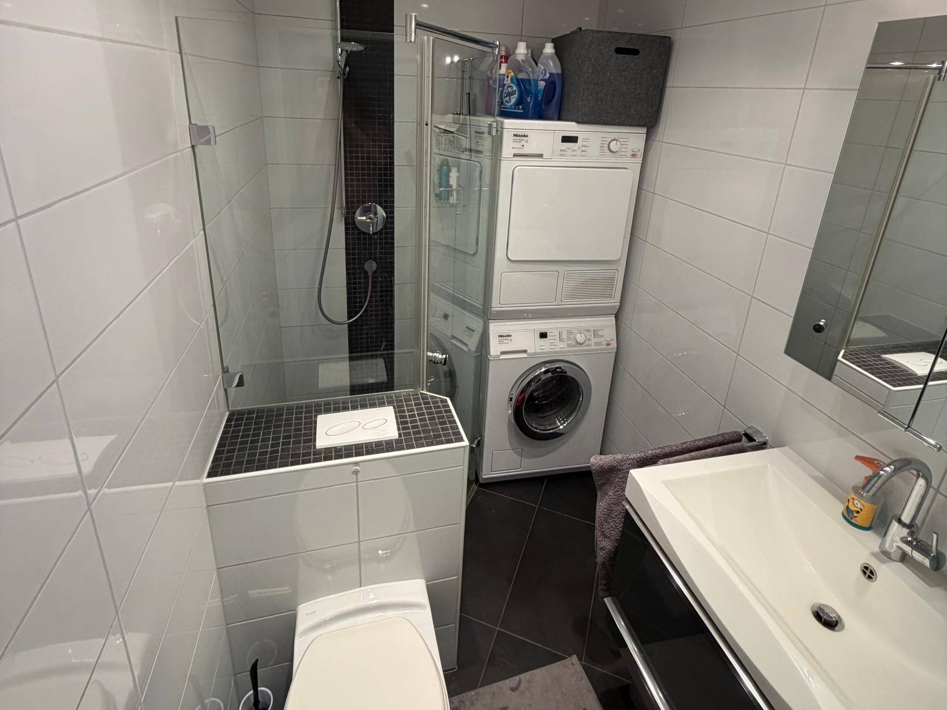 Modernes Badezimmer mit Dusche, Waschmaschine und Trockner sowie weißen Fliesen.