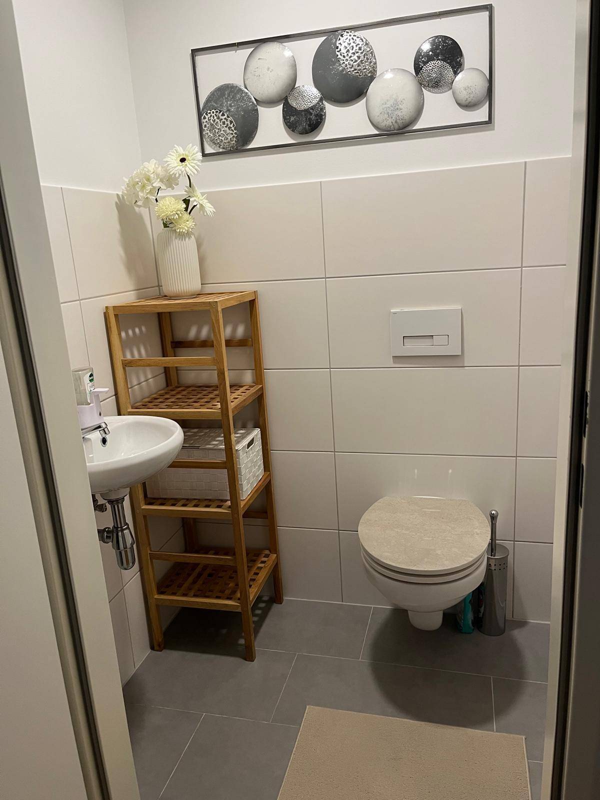 Kleines Gäste-WC mit Wand-WC, Waschbecken und Holzregal, funktional und hell gefliest.