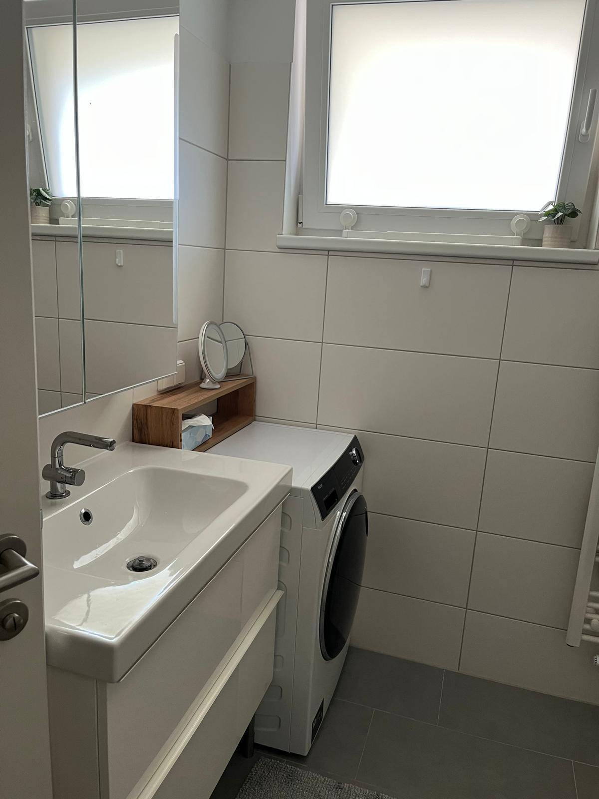 Helles Badezimmer mit Waschbecken, Spiegelschrank, Waschmaschine und Fenster für Tageslicht.