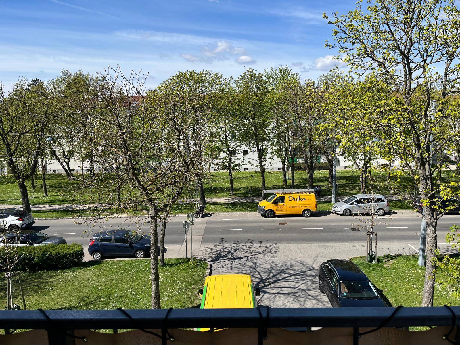 Ausblick vom Balkon auf eine grüne Straße mit Bäumen und vorbeifahrenden Fahrzeugen.