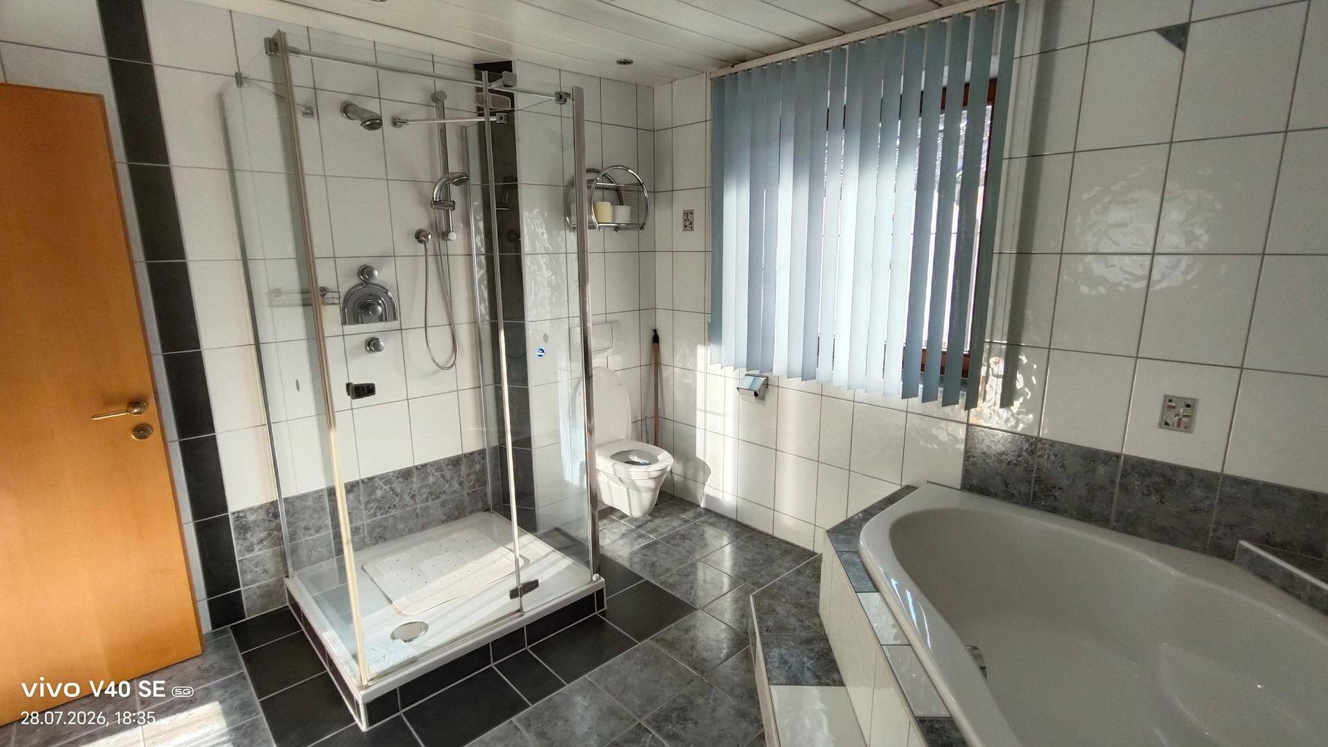 Helles Badezimmer mit Duschkabine, Badewanne, WC und Fenster mit Jalousien.