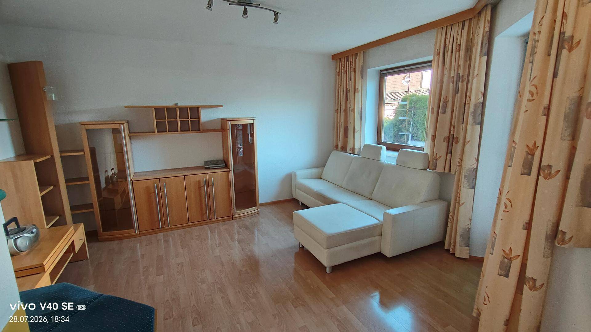 Komfortables Wohnzimmer mit hellem Sofa, Holzmöbeln und Vorhängen vor dem Fenster.