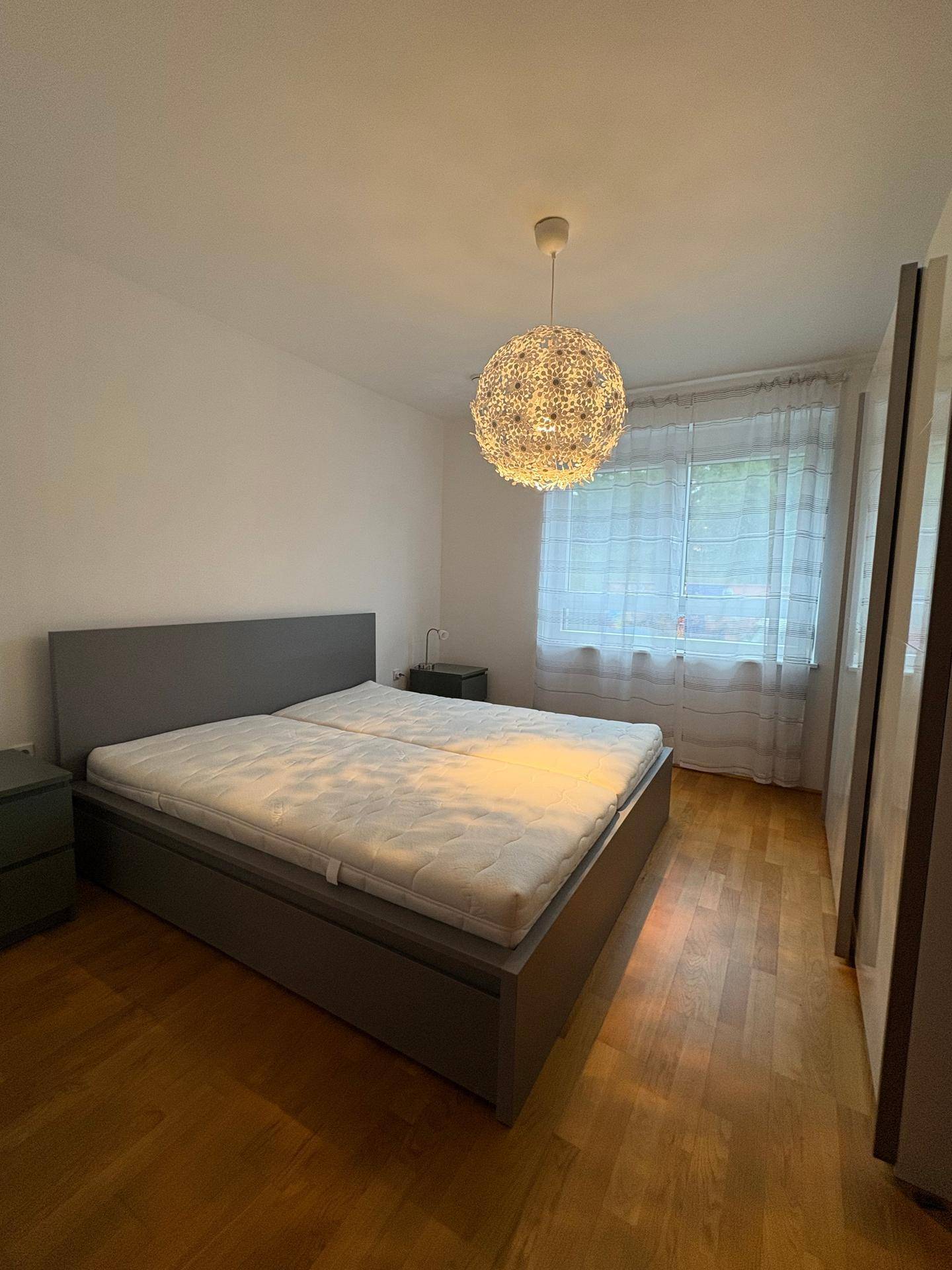 Schlafzimmer mit Doppelbett, Holzparkett und einem großen Fenster mit Vorhängen.