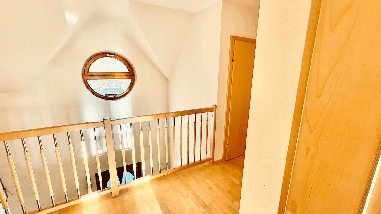 Heller Flur im Obergeschoss mit Holzgeländer und einem runden Fenster, das viel Licht hereinlässt.