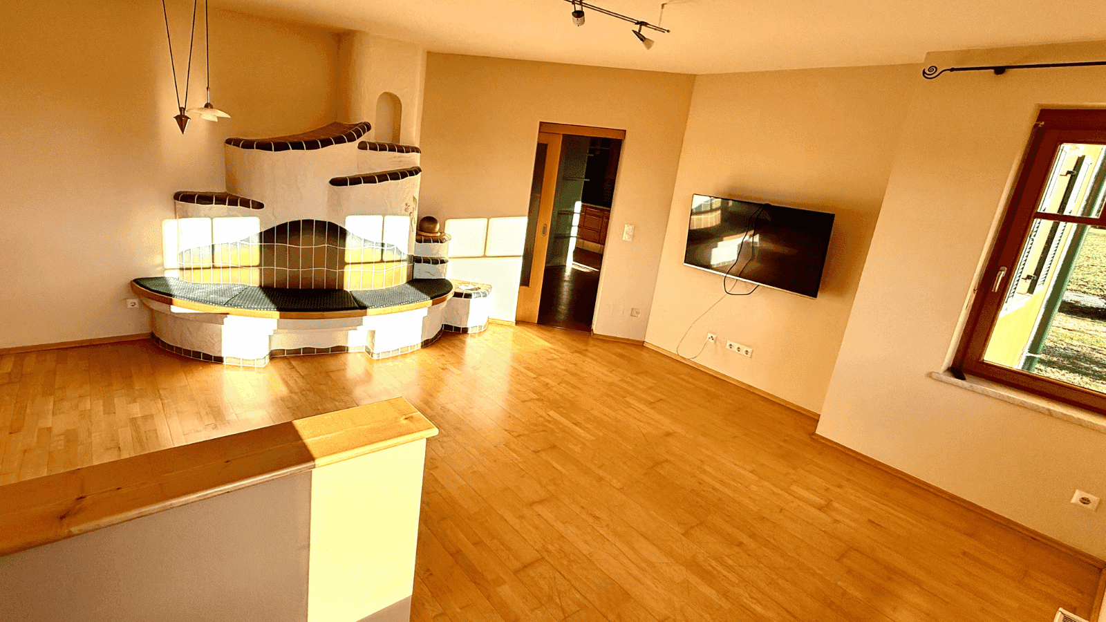 Geräumiges Wohnzimmer mit Holzboden, Kachelofen und einem Wandfernseher. Viel Sonnenlicht fällt durch die Fenster.