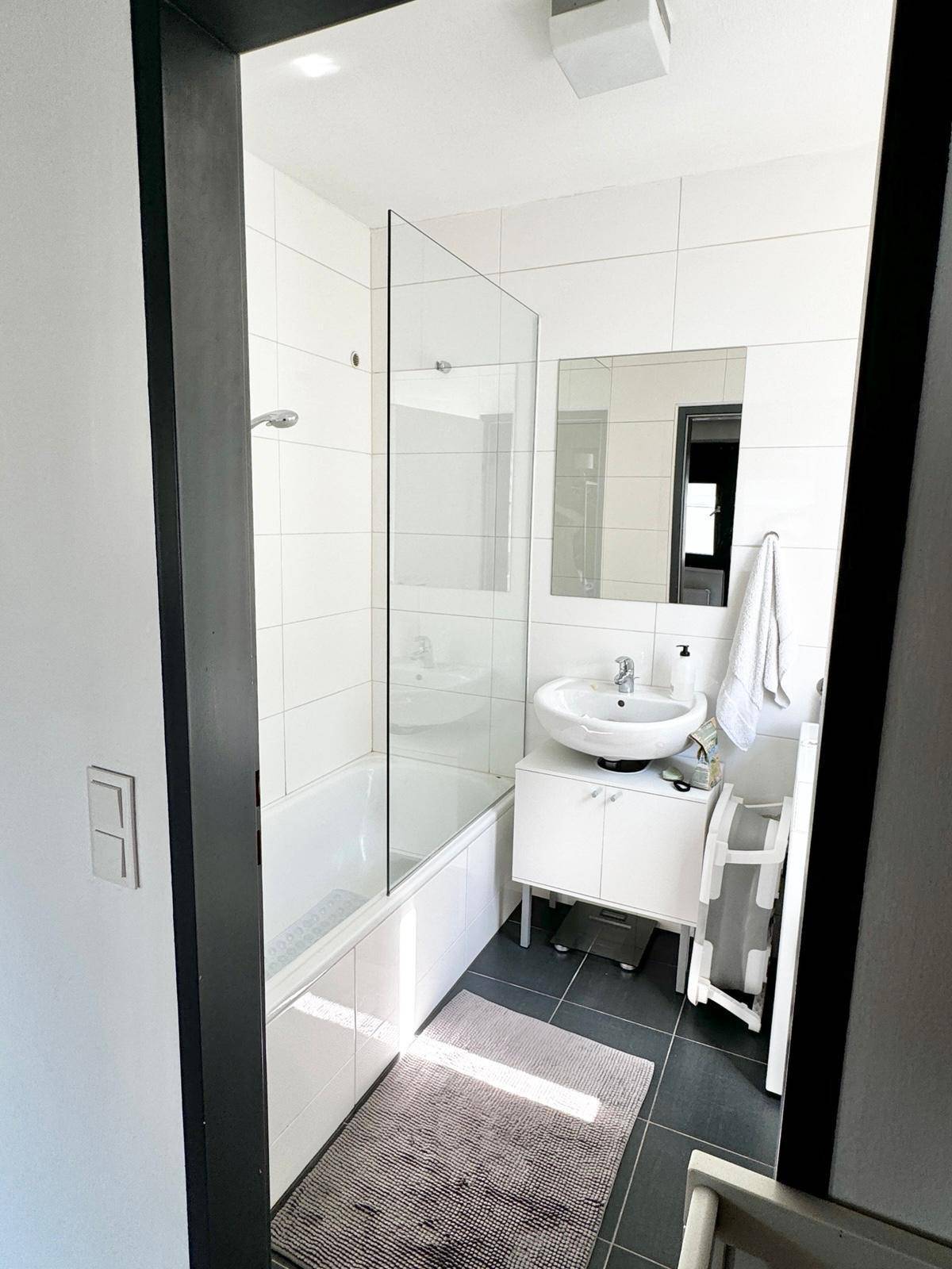 Modernes Badezimmer mit weißen Fliesen, einer Badewanne mit Duschwand und dunklem Fliesenboden.