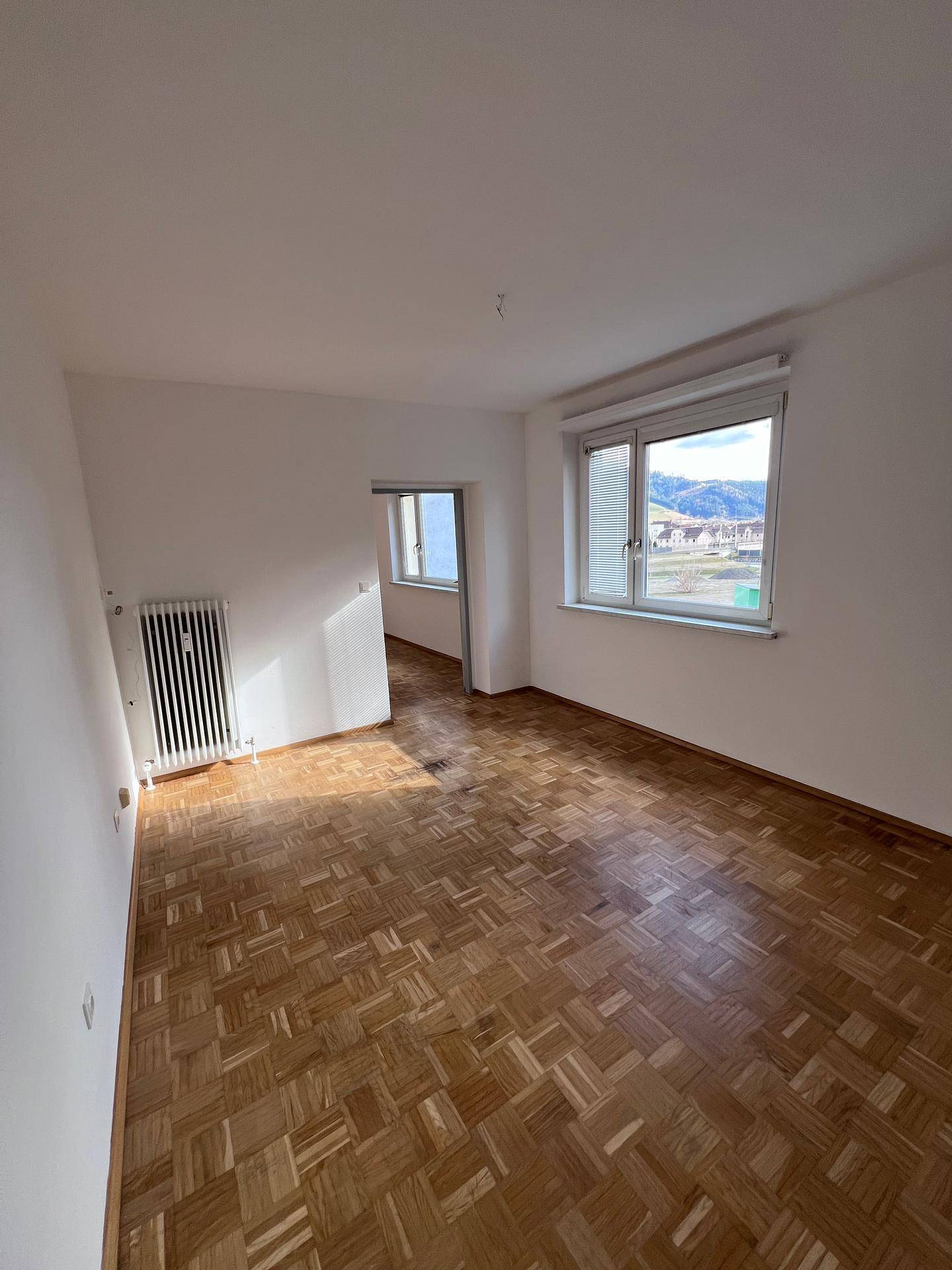 Geräumiges Zimmer mit Parkettboden und Heizkörper, das in einen weiteren Raum führt und hell ist.