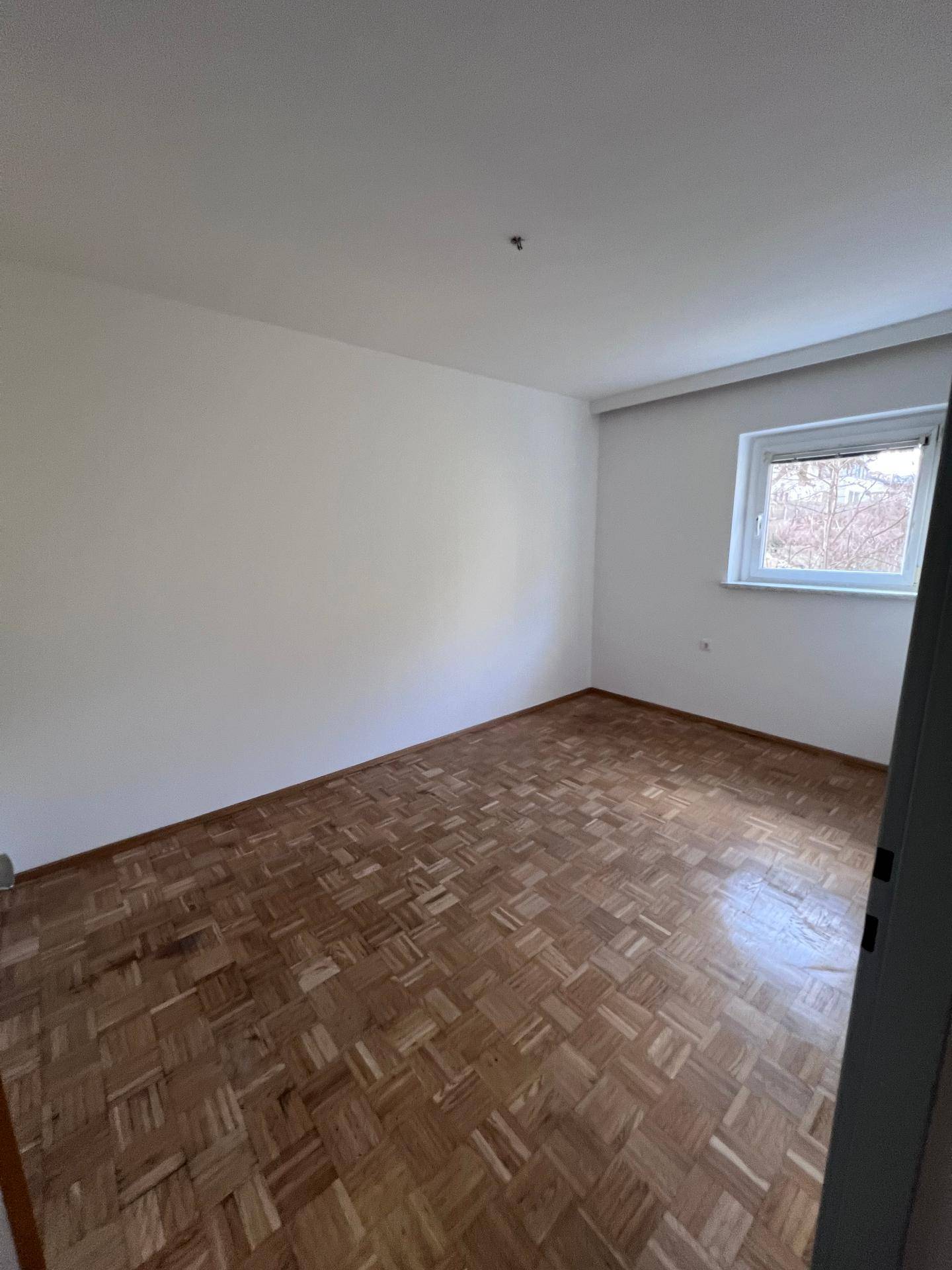 Helles Schlafzimmer mit Parkettboden und einem Fenster, das eine ruhige Aussicht auf Bäume bietet.