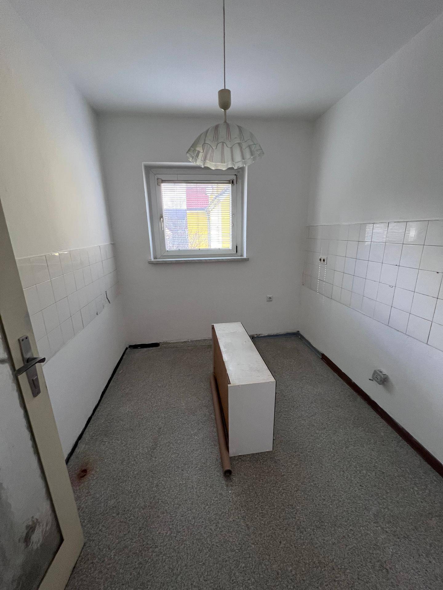 Leere Küche mit Fliesenboden, weißen Wandfliesen und einem Fenster mit Jalousien, bereit zur Gestaltung.