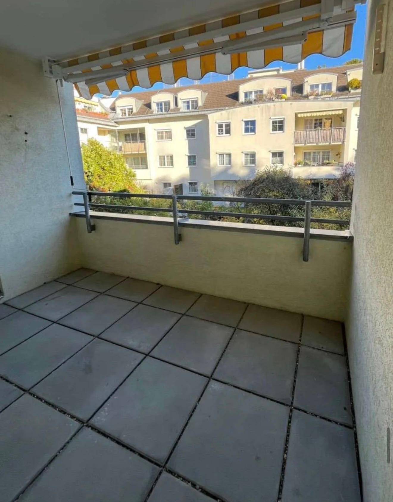 Balkon mit grauen Fliesen, gelb-weiß gestreifter Markise und Blick auf umliegende Gebäude.
