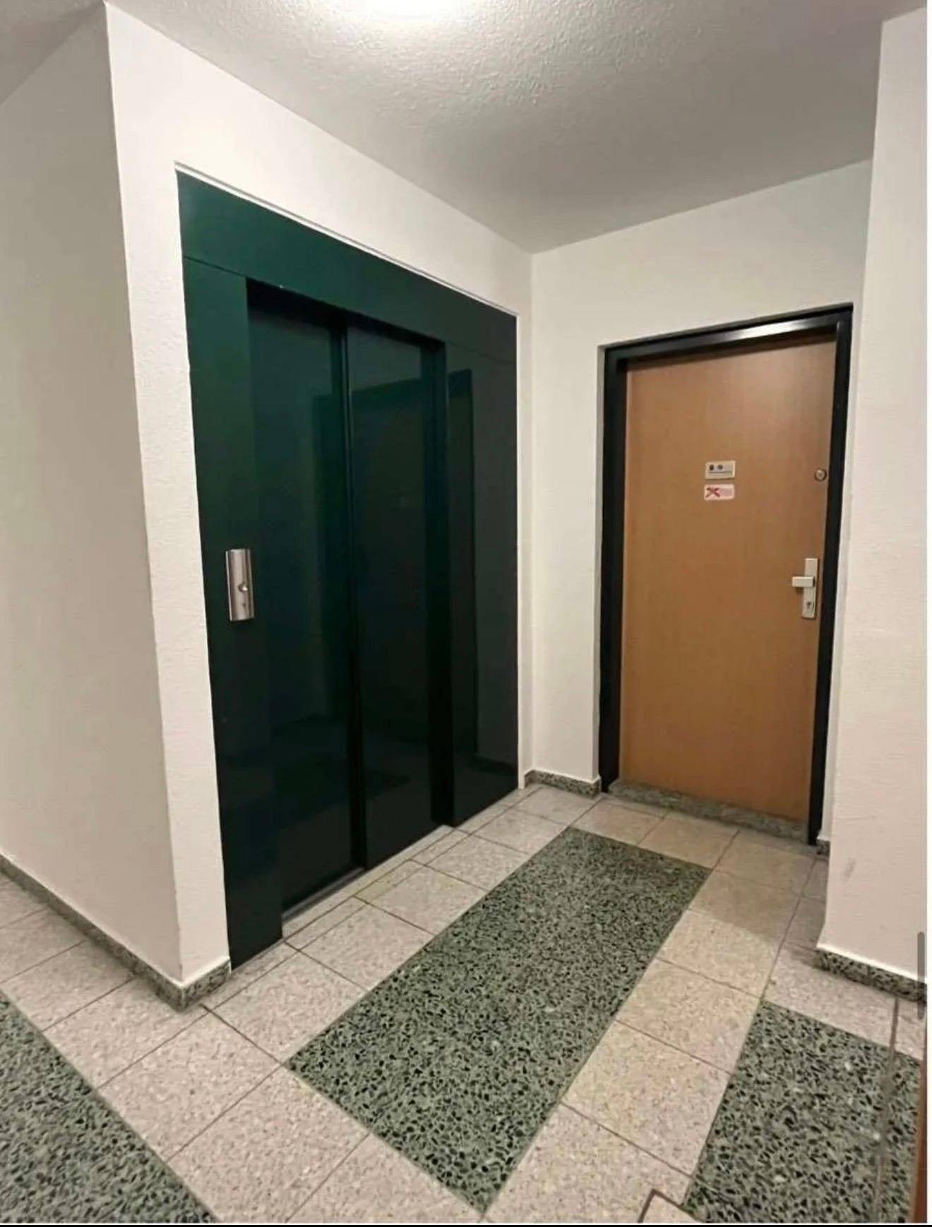Heller Hausflur mit grünem Aufzug und einer Wohnungstür.