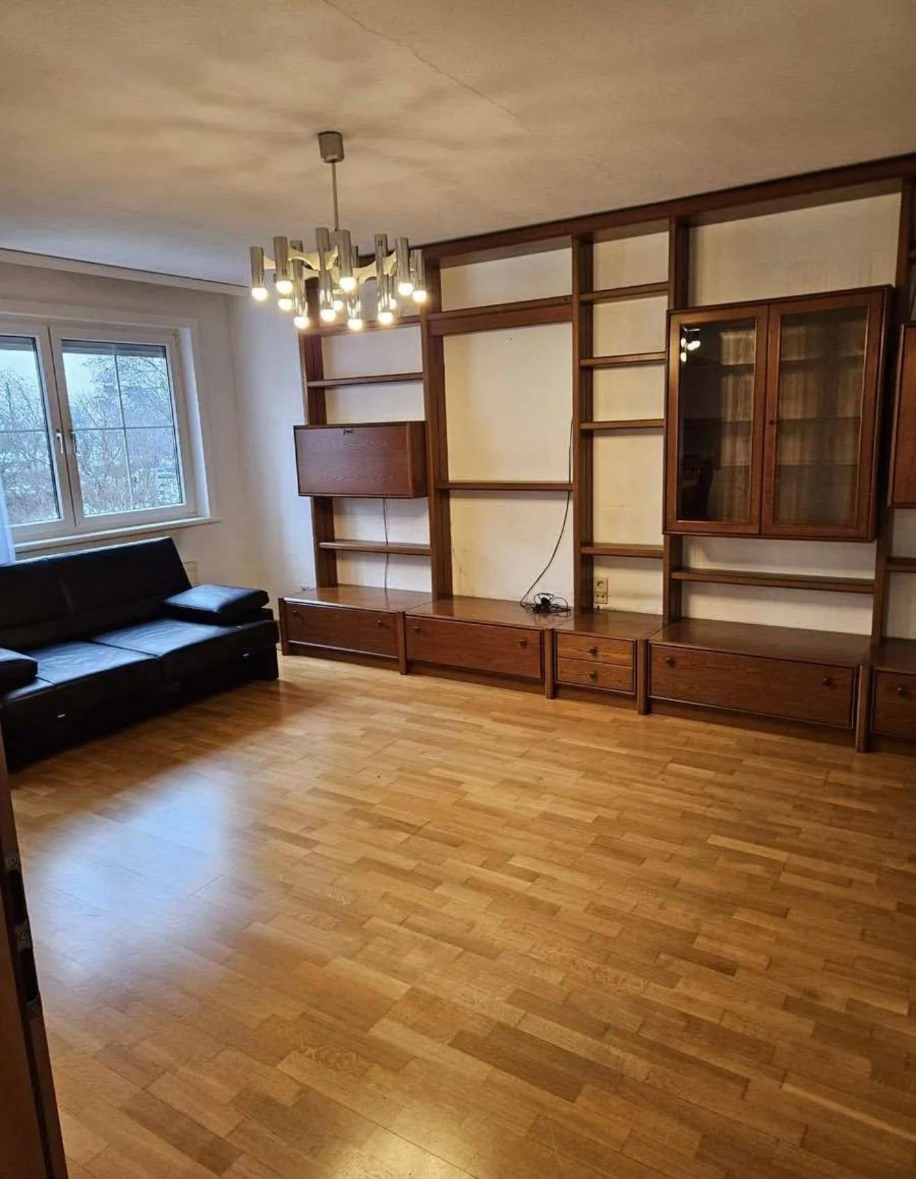 Geräumiges Wohnzimmer mit Parkettboden, großer Holzschrankwand und schwarzem Sofa.