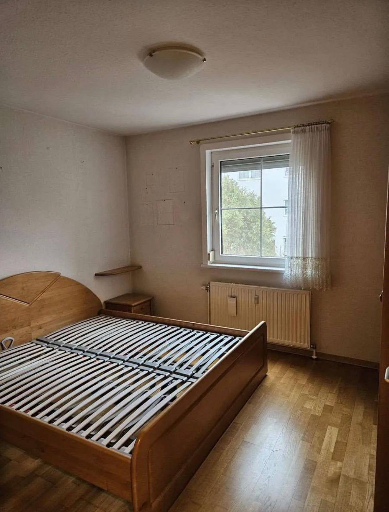 Schlafzimmer mit Holzbettrahmen, Parkettboden und einem Fenster mit Blick ins Grüne.