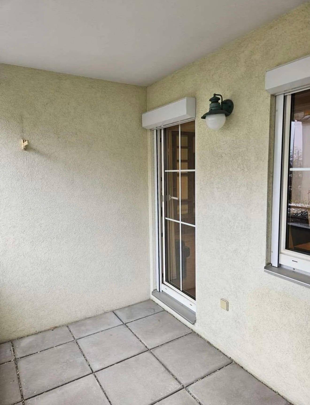 Gedeckter Balkon mit grauen Fliesen, einer Tür und einem Fenster mit Rollladen.