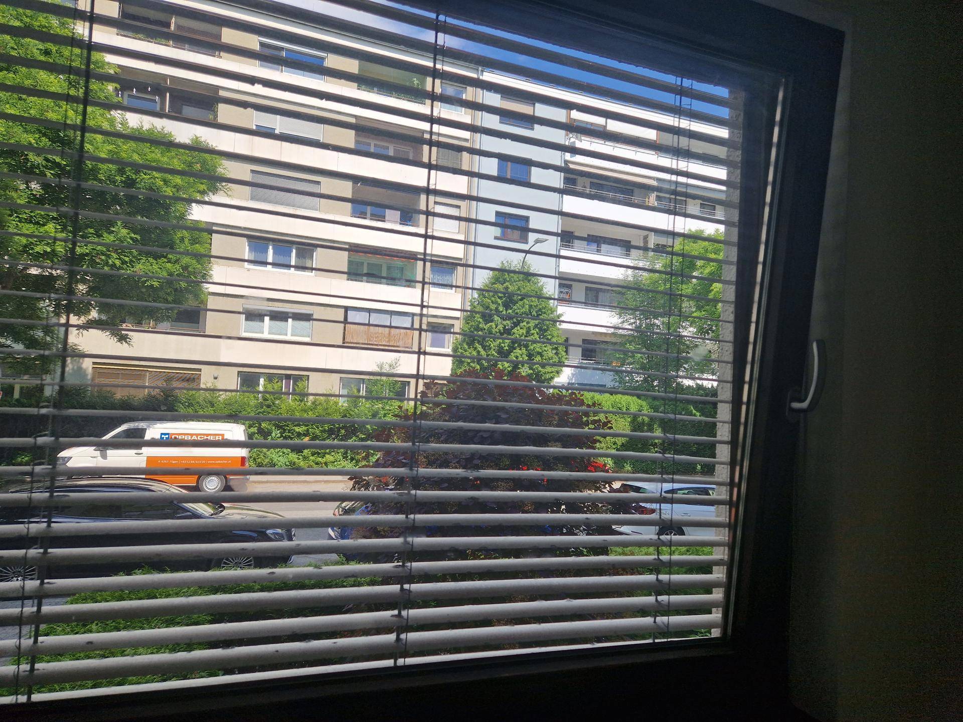Blick aus dem Fenster mit Jalousien auf die Straße und gegenüberliegende Gebäude.